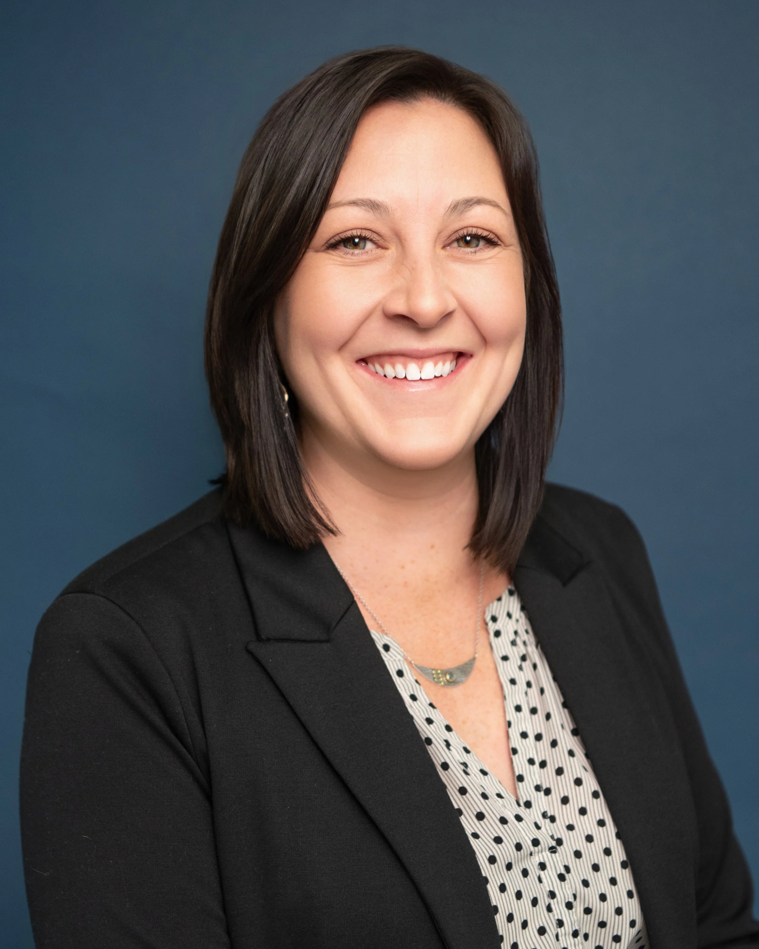 A smiling woman with dark brown hair styled in a bob, wearing a black blazer over a white blouse with black polka dots and a layered necklace, against a solid blue background.