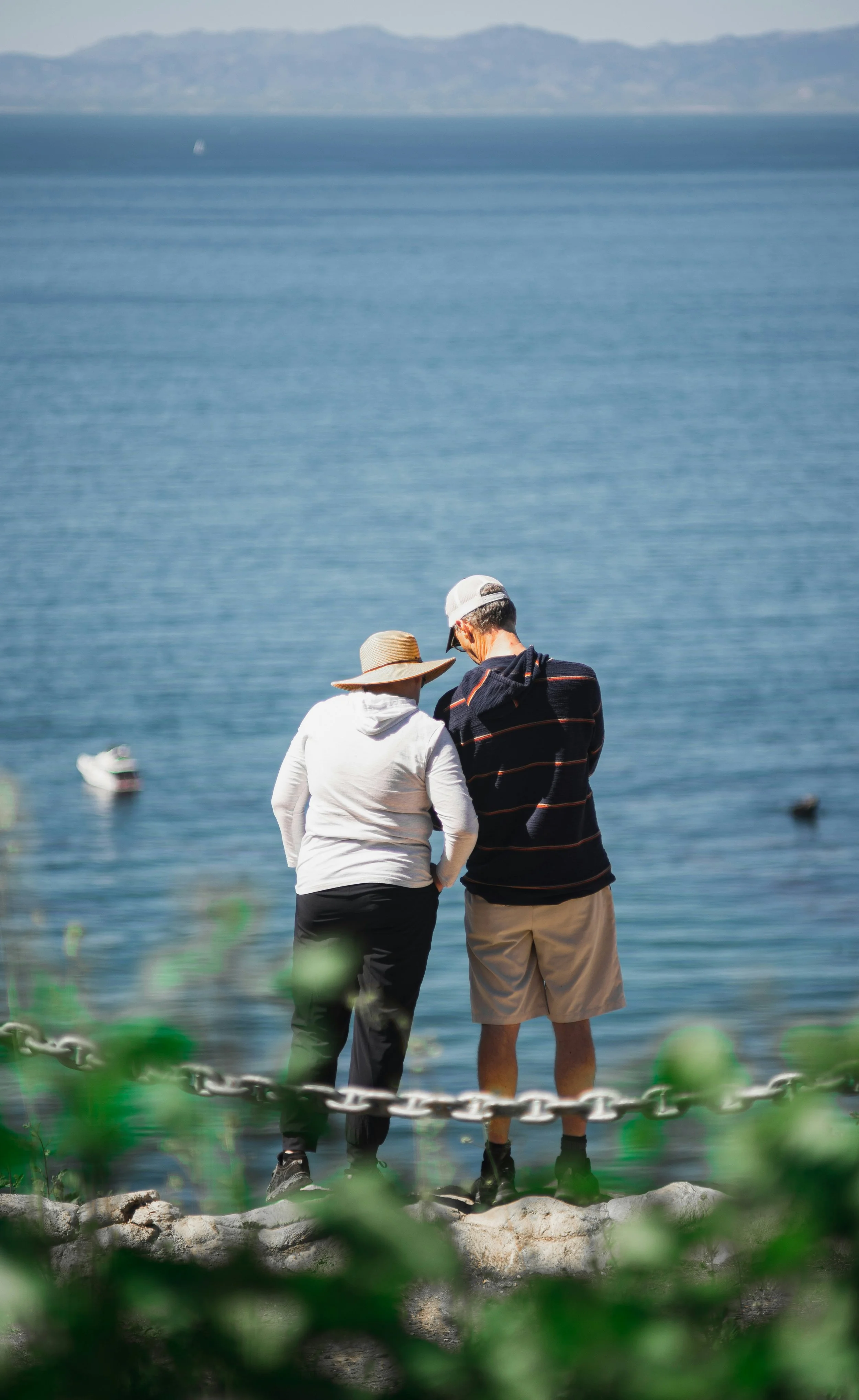 Two people standing on rocks by the water's edge, facing the ocean, with sailboats in the distance and mountains in the background.