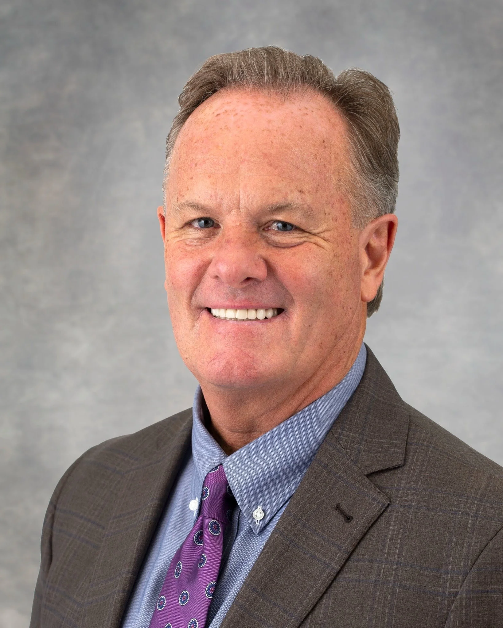 Close-up of a smiling middle-aged man with gray hair, wearing a brown suit, a purple tie with circular patterns, and a light blue shirt, against a gray background.