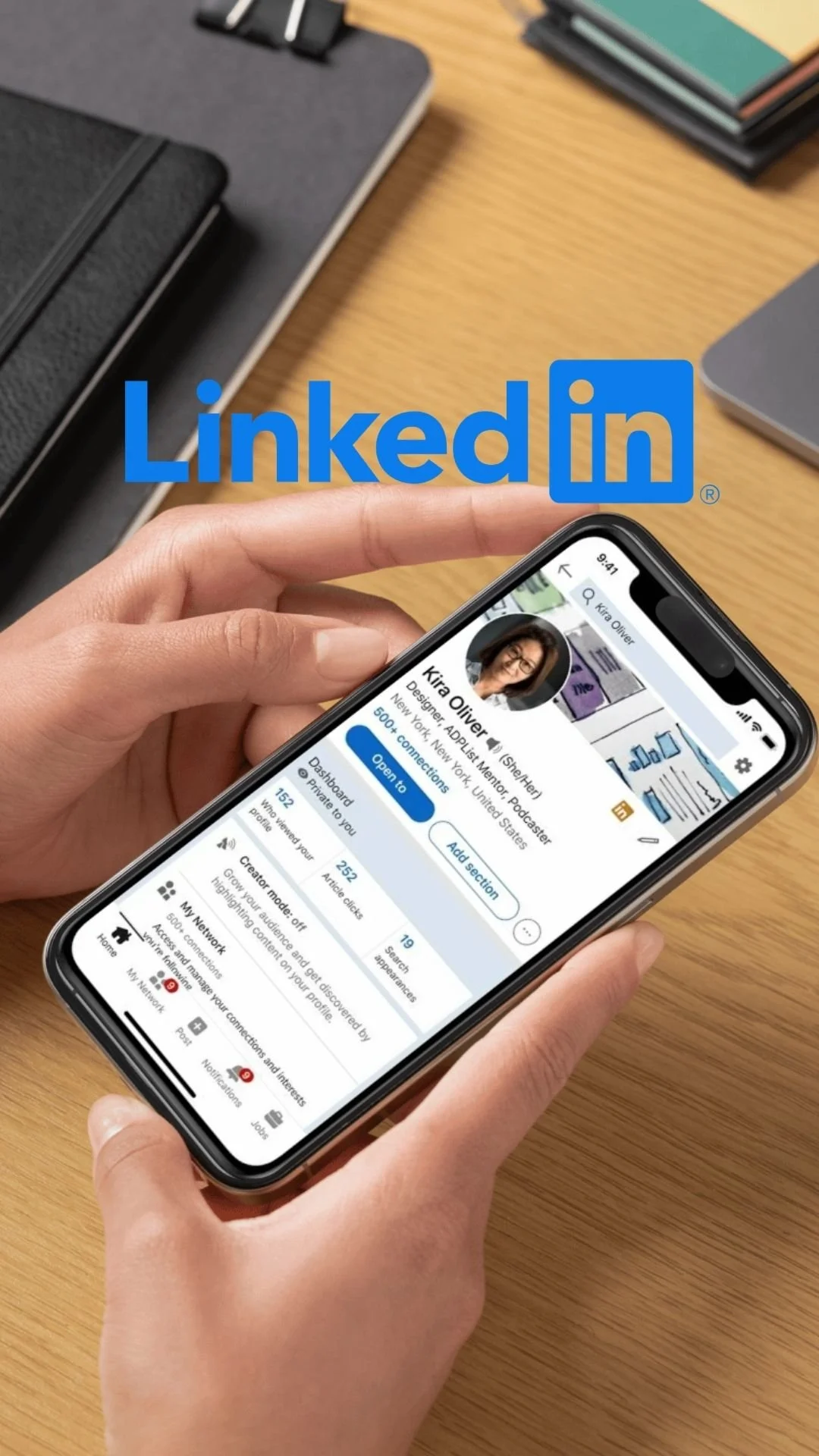 Close-up of a person's hand holding a smartphone displaying a LinkedIn profile of Kira Oliver, with office supplies and a wooden desk in the background.