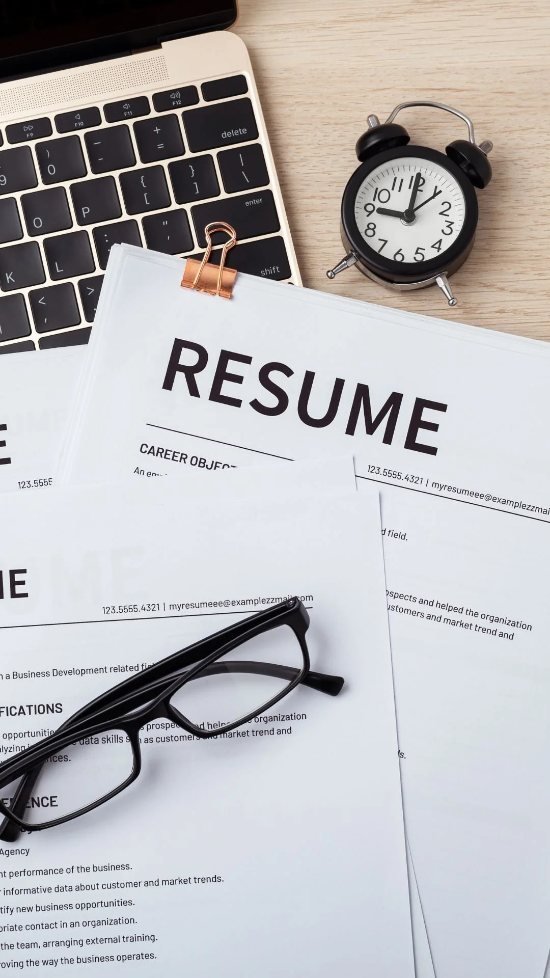 A desk setup with a laptop, a black alarm clock showing 12:00, a stack of resumes with a pair of eyeglasses on top, and a pair of glasses resting on the table.