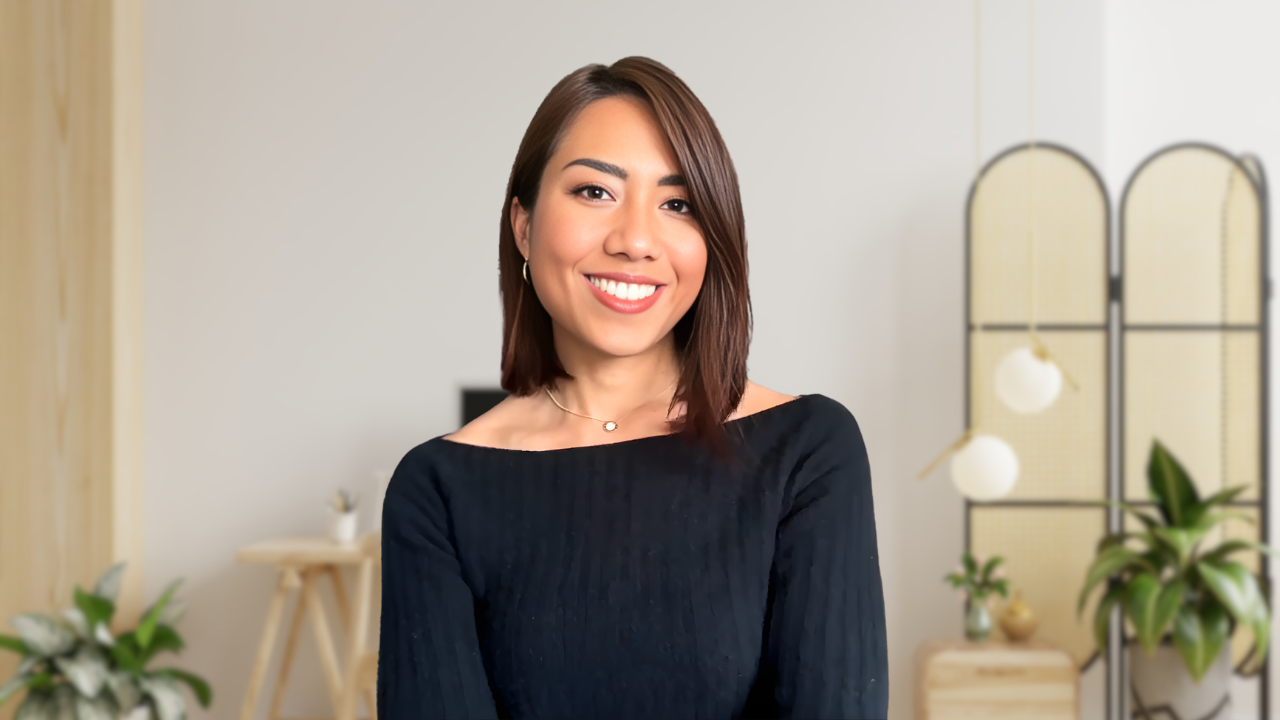 A woman with shoulder-length brown hair smiling, wearing a black top, standing in a bright, modern interior with plants and decor.