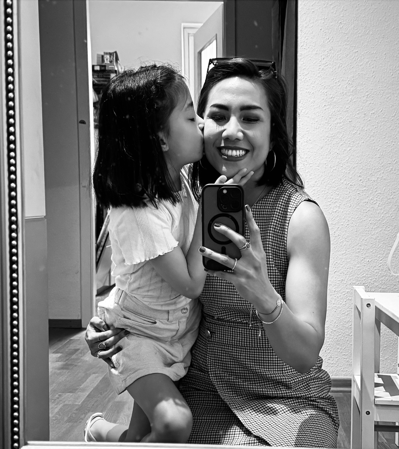 A woman taking a mirror selfie with a young girl giving her a kiss on the cheek. The woman is smiling with glasses on her head, wearing a sleeveless checkered dress. The girl is wearing a light-colored top and shorts, and has her eyes closed as she kisses the woman.