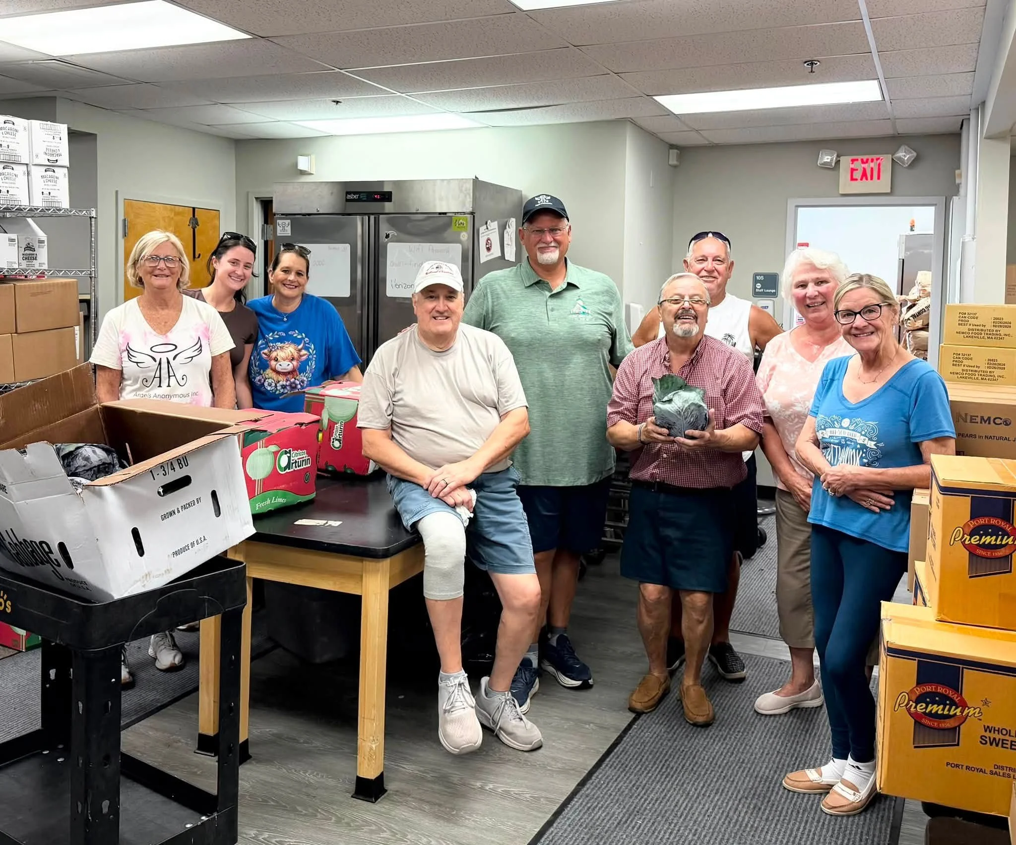 A group of nine people, including men and women of various ages, standing and smiling inside a room with shelves and boxes, possibly in a food bank or donation center.