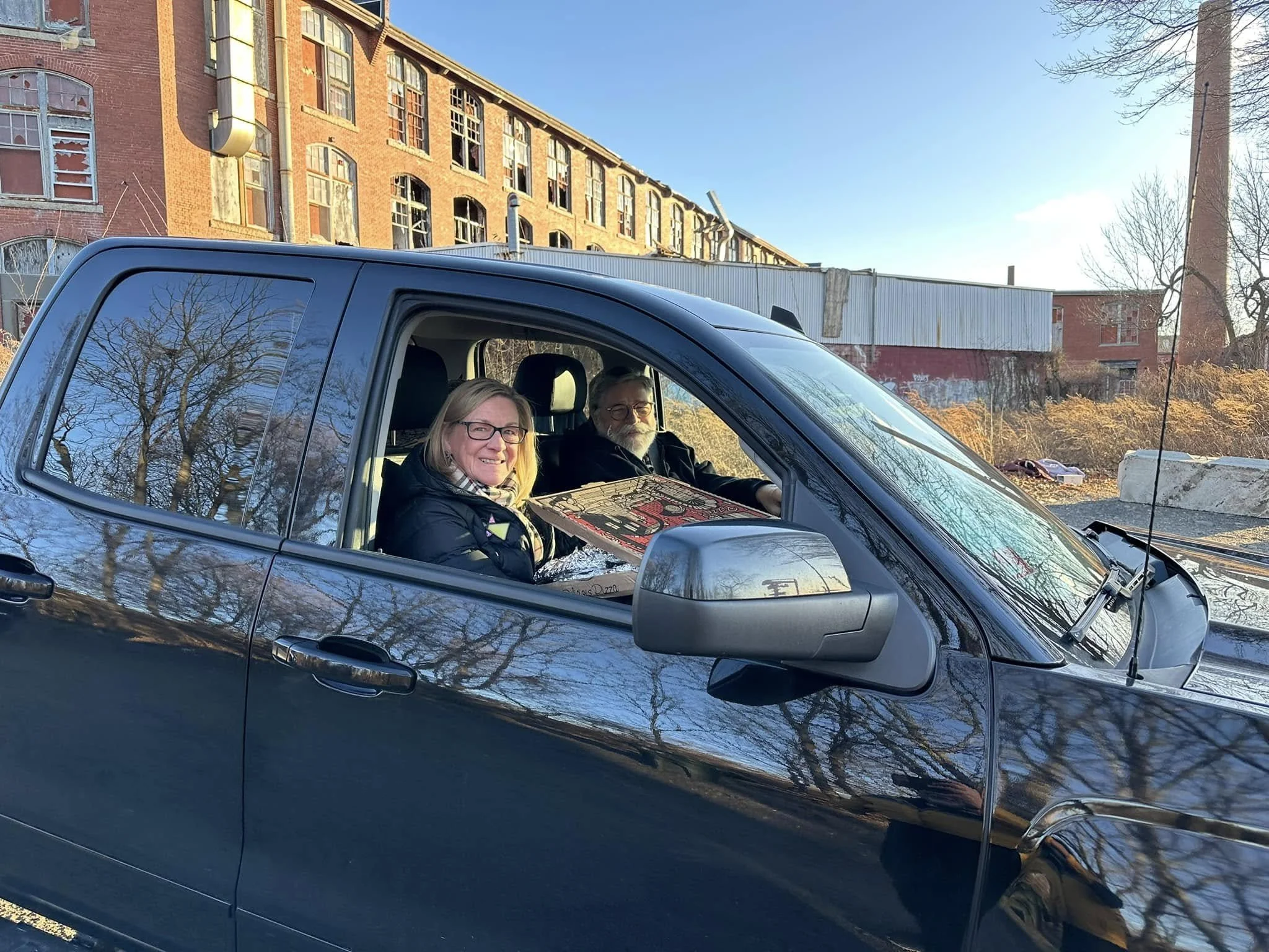 A woman and a man sitting inside a dark-colored pickup truck parked outdoors. The woman is smiling and wearing glasses and a black jacket, while the man has a beard and glasses, and he is holding a pizza box. There are old brick buildings with broken