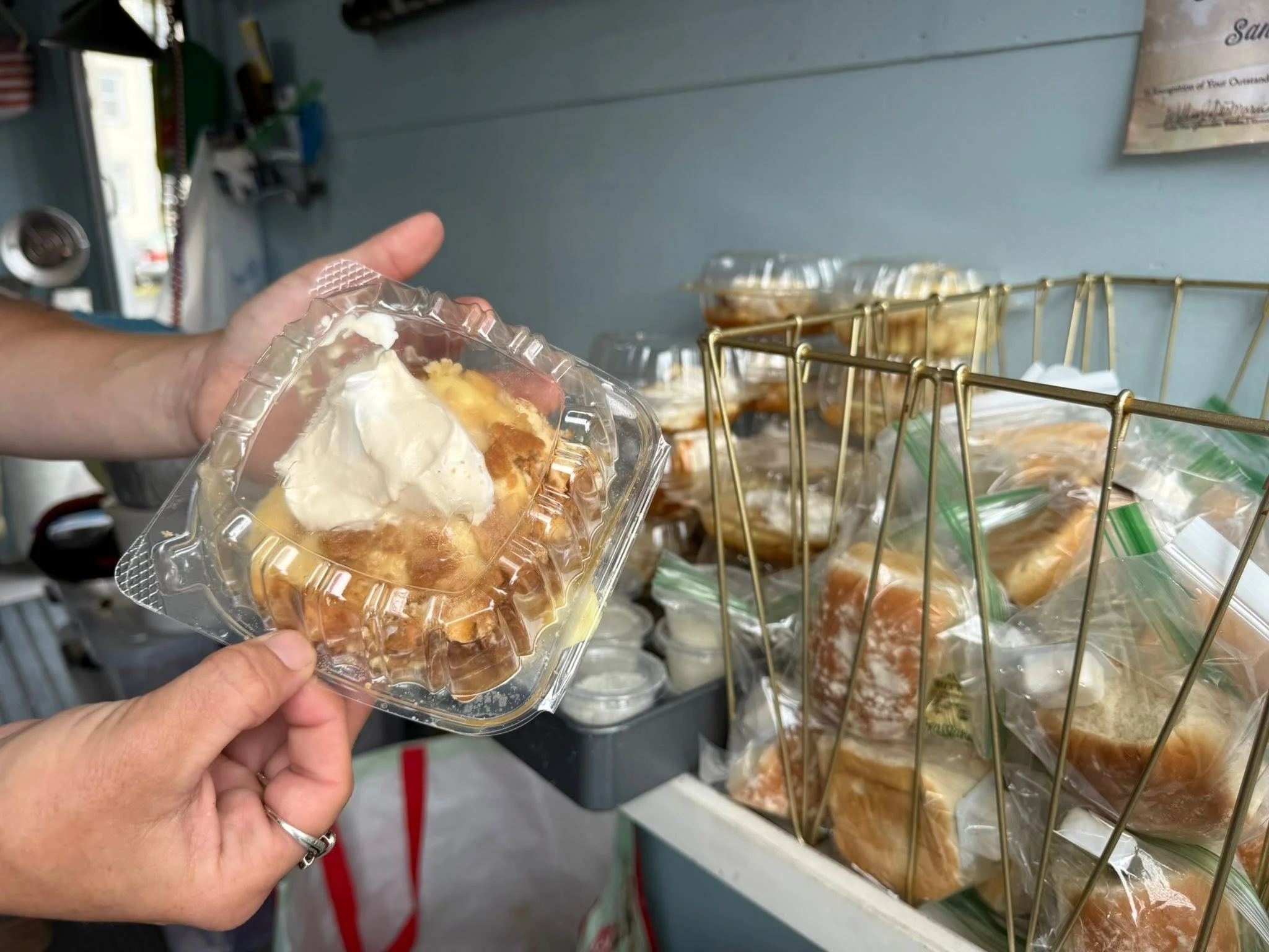 A person holding a clear plastic container with a dessert topped with whipped cream and caramel sauce. In the background, there are shelves with pre-packaged desserts in clear containers.
