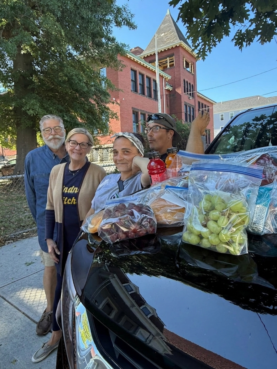 Four people stand around a black car filled with plastic bags of grapes and other snacks. They are outdoors on a sidewalk in front of a red brick building with a tower. The group appears to be gathering for a casual occasion, with trees and a clear b