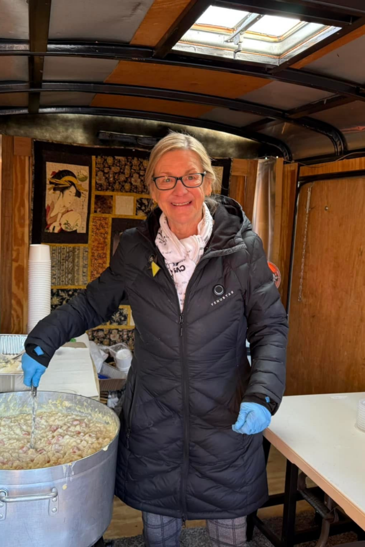 An elderly woman with glasses, blonde hair, and a black jacket is smiling while stirring a large pot of food. She is wearing blue gloves and standing inside a wooden structure with a decorative Japanese-style wall hanging in the background. There are