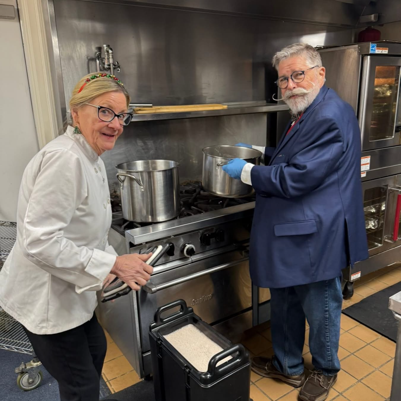 An elderly woman in a chef's coat and glasses, and an elderly man in a blue jacket with glasses and a mustache, cooking together in a professional kitchen, stirring pots on a stove.