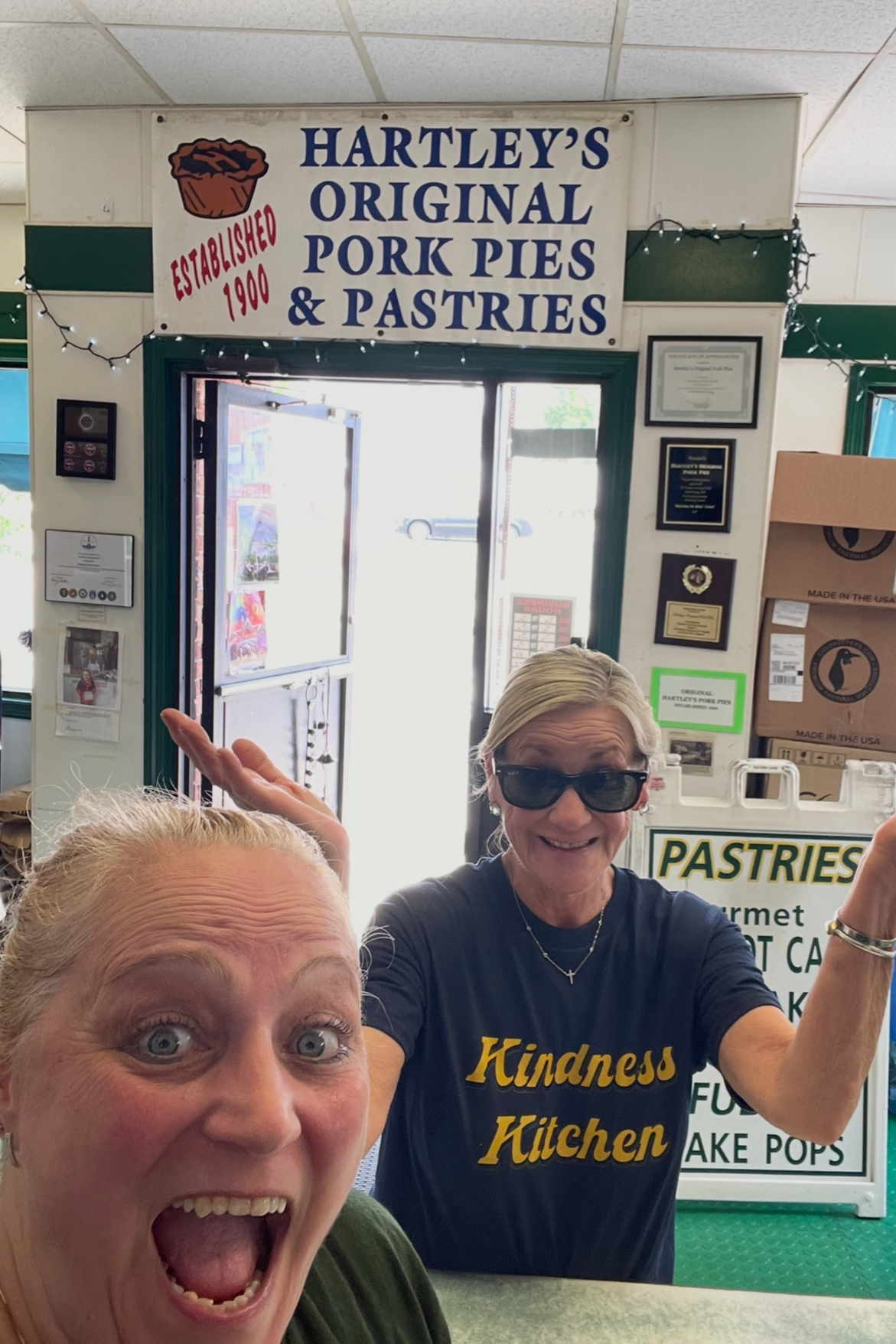 Two women inside Hartley's restaurant, one in the foreground with a surprised expression and the other in the background wearing sunglasses, smiling; sign above reads "Hartley's Original Pork Pies & Pastries."