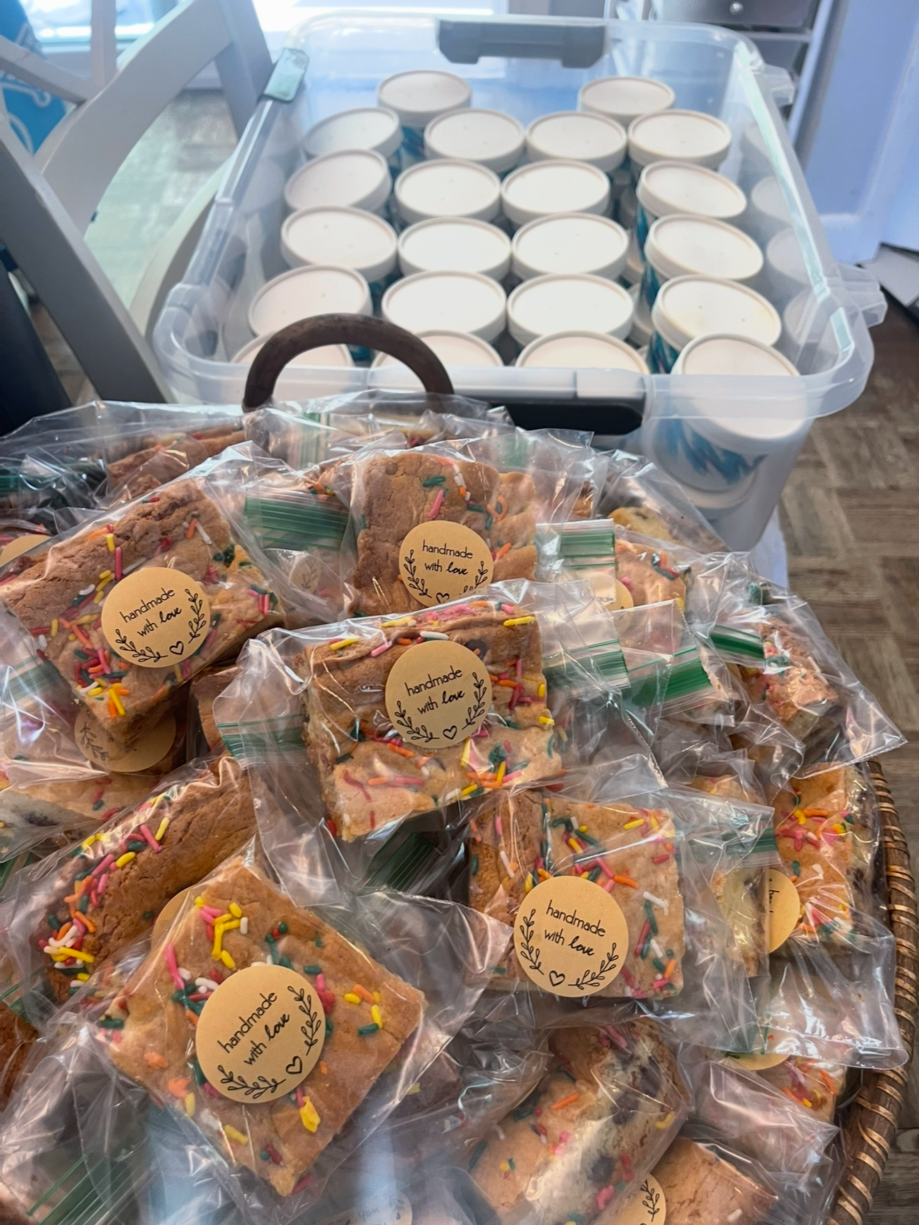 A large plastic container filled with white yogurt cups and a basket of individually wrapped homemade cookies with colorful sprinkles, each cookie labeled 'handmade with love'.