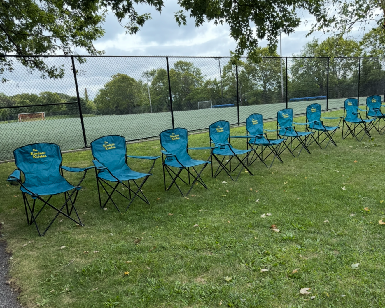 Blue folding chairs with yellow text reading 'The Kiwanis Kitchen' lined up on green grass next to a fenced sports field under a cloudy sky.