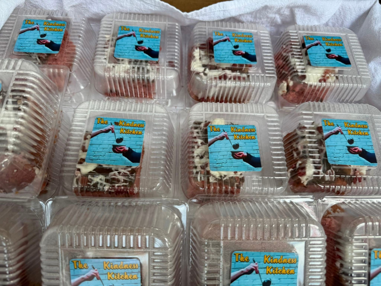 Multiple clear plastic containers with cakes inside, each labeled with a sticker that says "The Kindness Kitchen" featuring an illustration of a hand holding a spoon over a blue brick background.