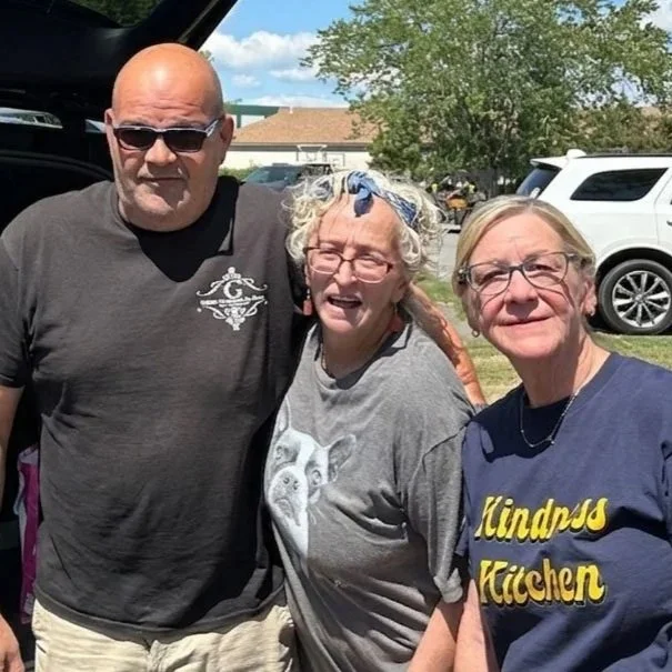 Three middle-aged people outdoors near parked cars, smiling at the camera on a sunny day.