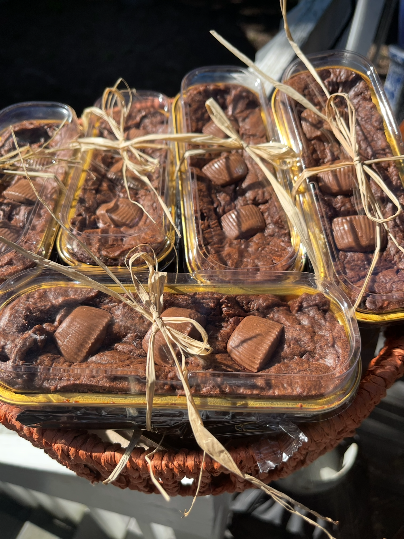 Multiple small plastic containers filled with homemade chocolate brownies topped with pieces of chocolate bar, tied with raffia bows, placed on a woven tray.