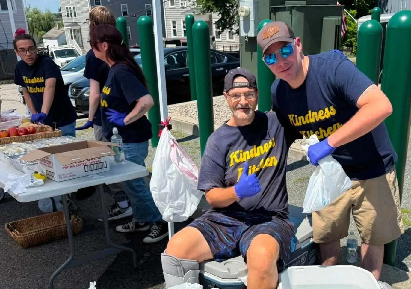 Group of volunteers wearing matching 'Kindness Kitchen' t-shirts, working together to distribute food, with apples and supplies on the table, outdoors on a sunny day.