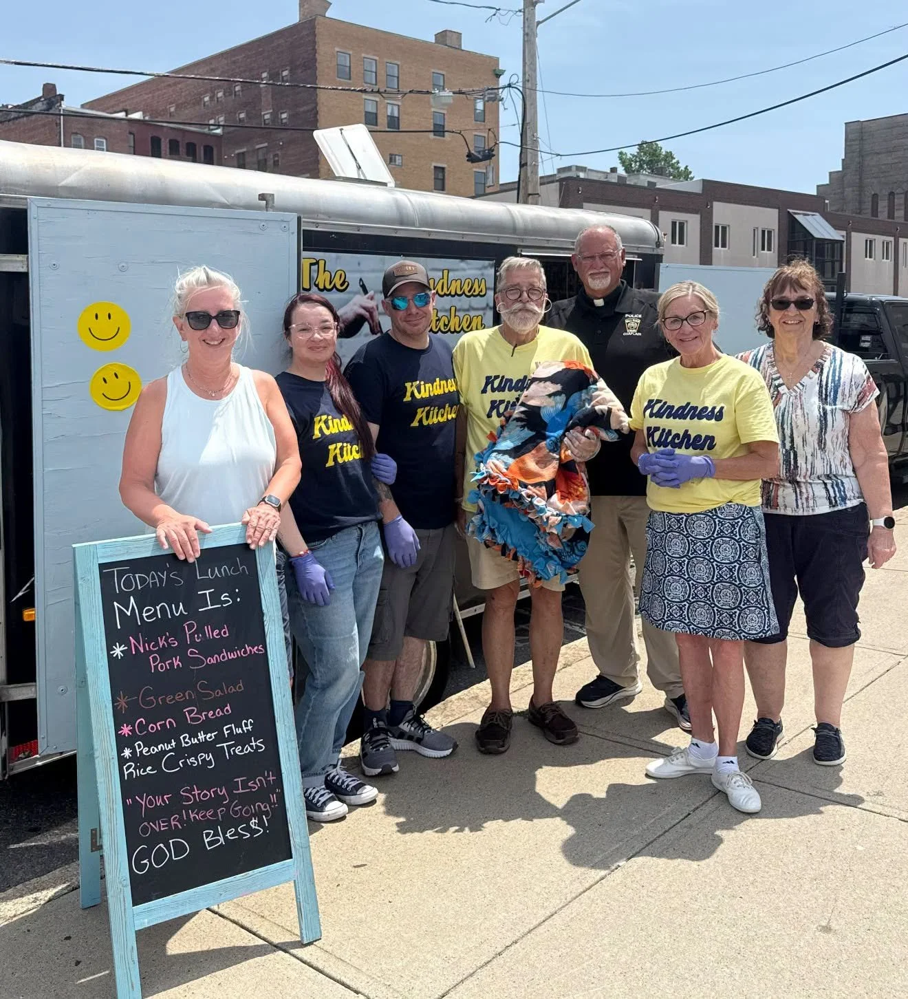 Group of people standing in front of a food truck with a menu board. The group includes five women and two men, some wearing shirts that say 'Kindness Kitchen.' The women are smiling, and some are wearing purple gloves. The food truck has a sign that says 'The Kindness Kitchen,' with two smiley face stickers. The menu board lists lunch options such as Nick's Pilled Pork Sandwiches, green salad, corn bread, peanut butter fluff rice crispy treats, and a note encouraging kindness.