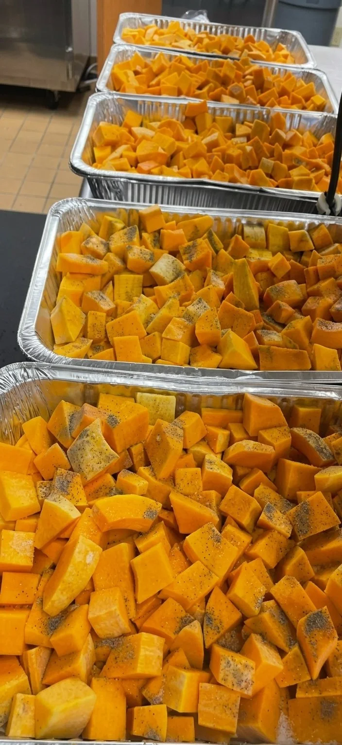 Multiple aluminum trays filled with cubed raw butternut squash, some with seasoning on top, placed on a kitchen countertop.