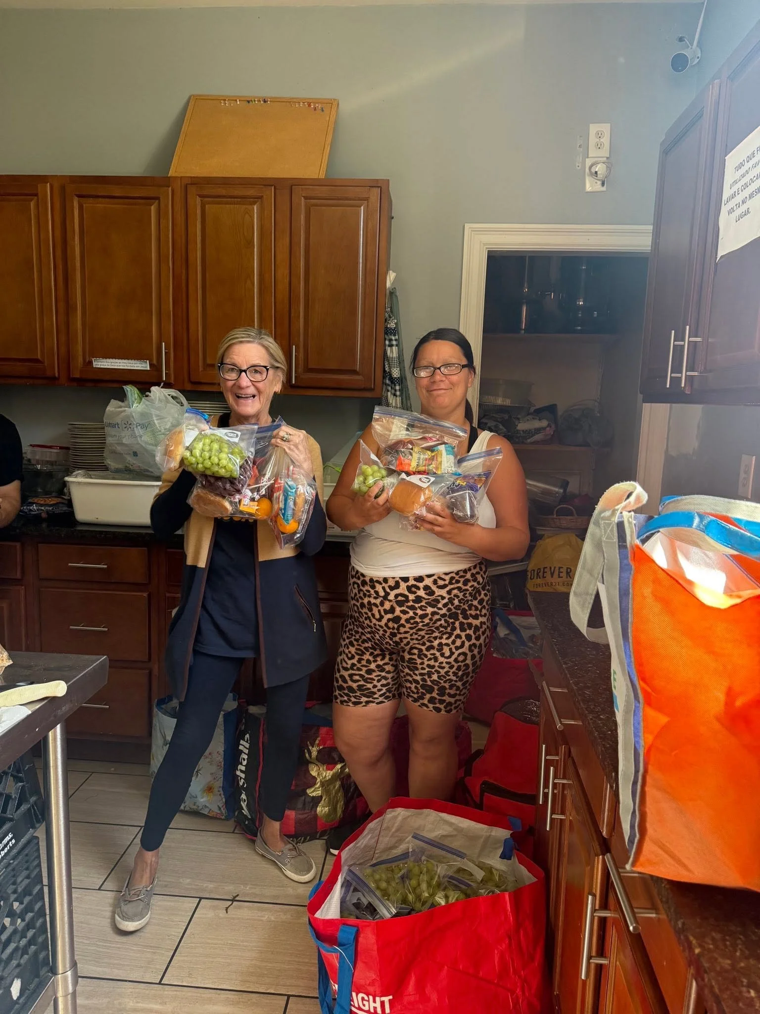 Two women smiling and holding bags of grapes and bread in a kitchen, surrounded by grocery bags and food items.