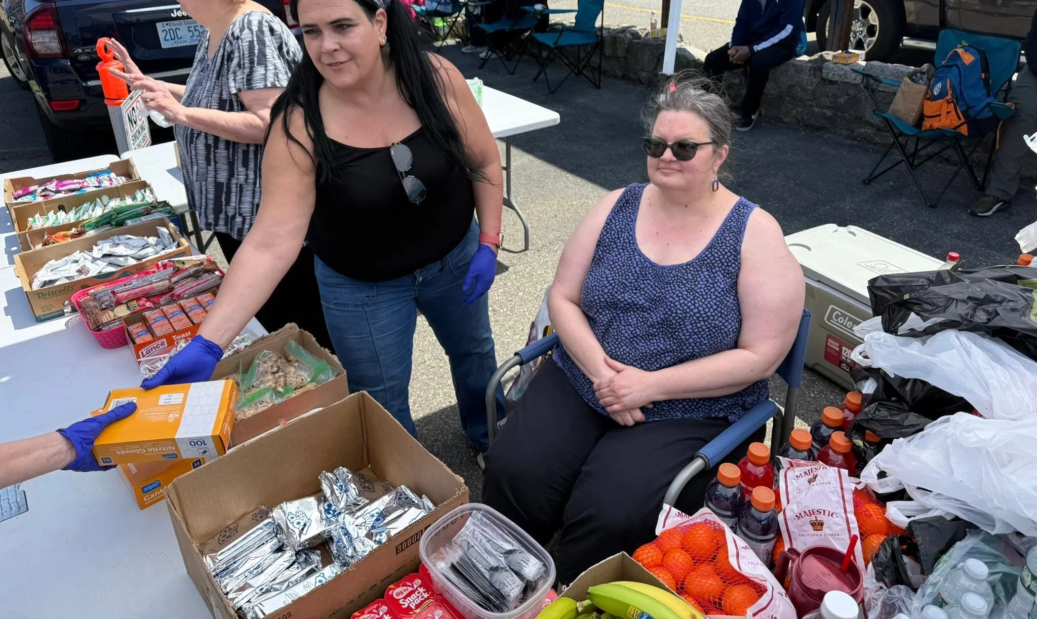 Women at a table organizing food donations outdoors, with boxes of snacks, drinks, and produce visible.