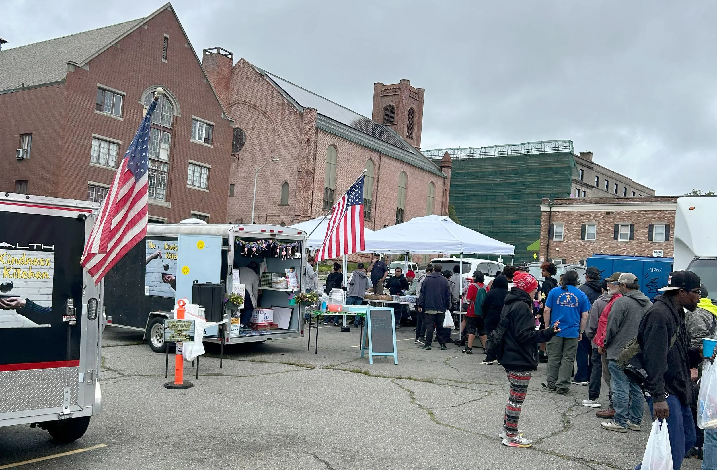 A street fair or market taking place in front of a large brick church with a tower. People are gathered around vendor stalls and food trucks, with American flags displayed. Overcast sky.