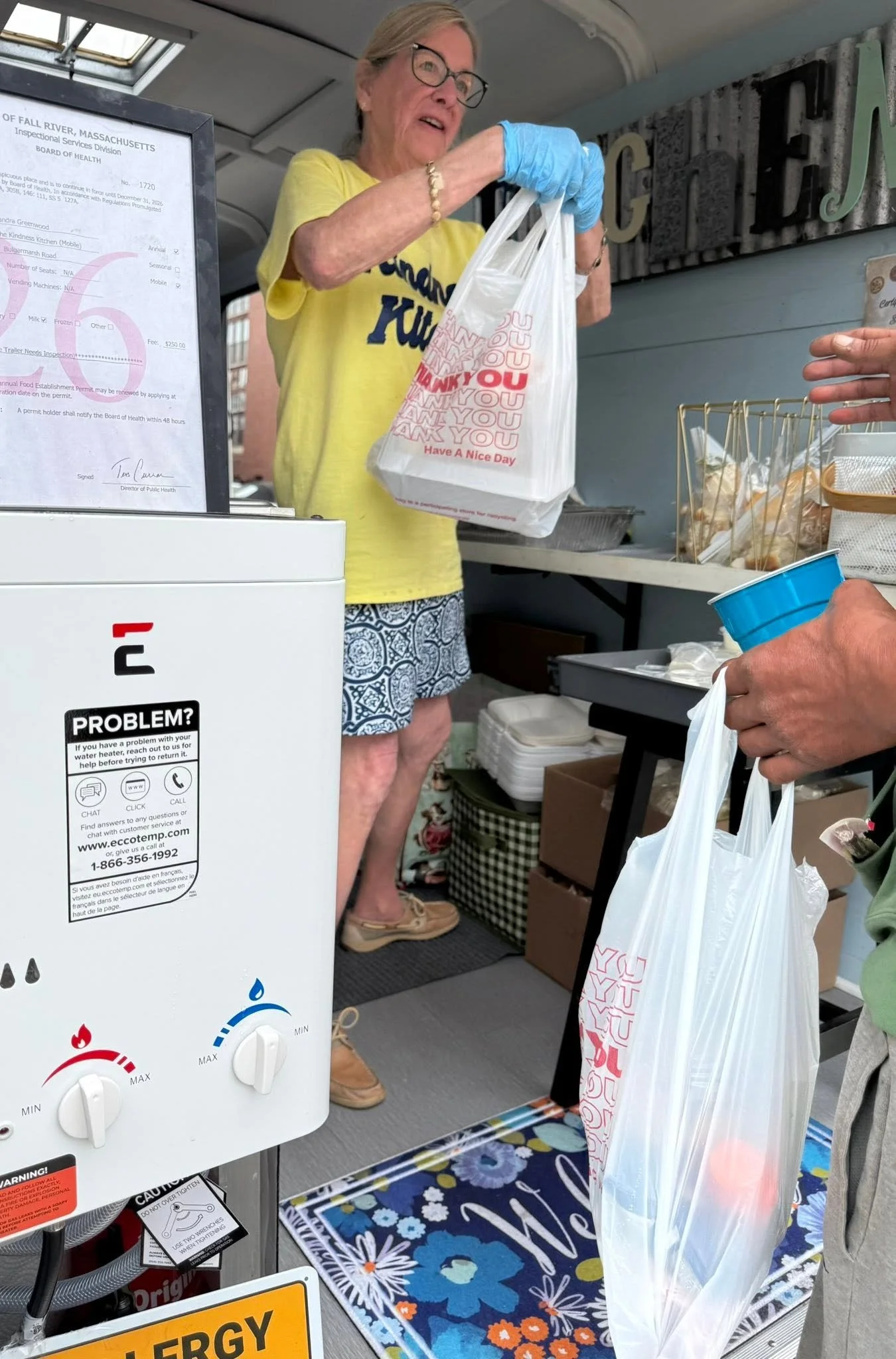 A woman wearing glasses, a yellow shirt, and blue gloves is handing a white plastic bag to another person. Behind her is a sign with a document, and in the foreground, part of a water heater and a floral rug are visible.