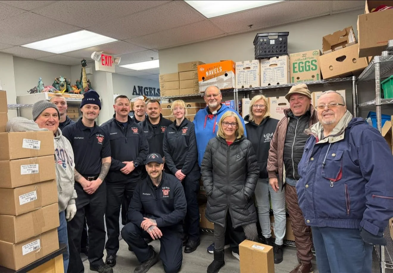 A group of people inside a storage room or warehouse, surrounded by boxes and shelves. Some are wearing dark uniforms, others are in casual winter clothing. They are smiling and posing for the photo.