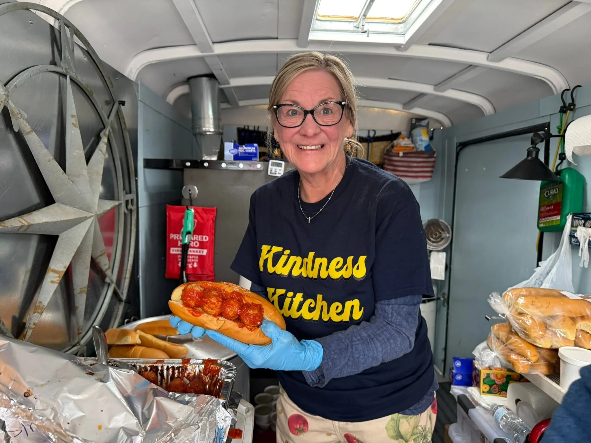 A woman with blonde hair, glasses, and wearing a navy blue shirt with yellow text, is smiling and holding a pepperoni pizza in a food truck or kitchen. She is wearing blue gloves, and the background includes various food items, kitchen equipment, and supplies.