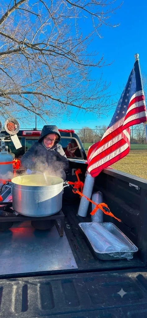 A woman in a hoodie stands in the back of a pickup truck, cooking a large pot of soup or stew outdoors. An American flag is attached to the truck bed. The scene is set on a clear and sunny day with leafless trees in the background.
