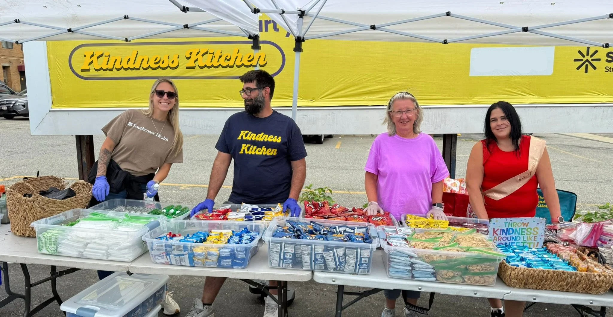 Four people standing behind a table filled with snacks at an outdoor event, with a yellow banner reading 'Kindness Kitchen' in the background.