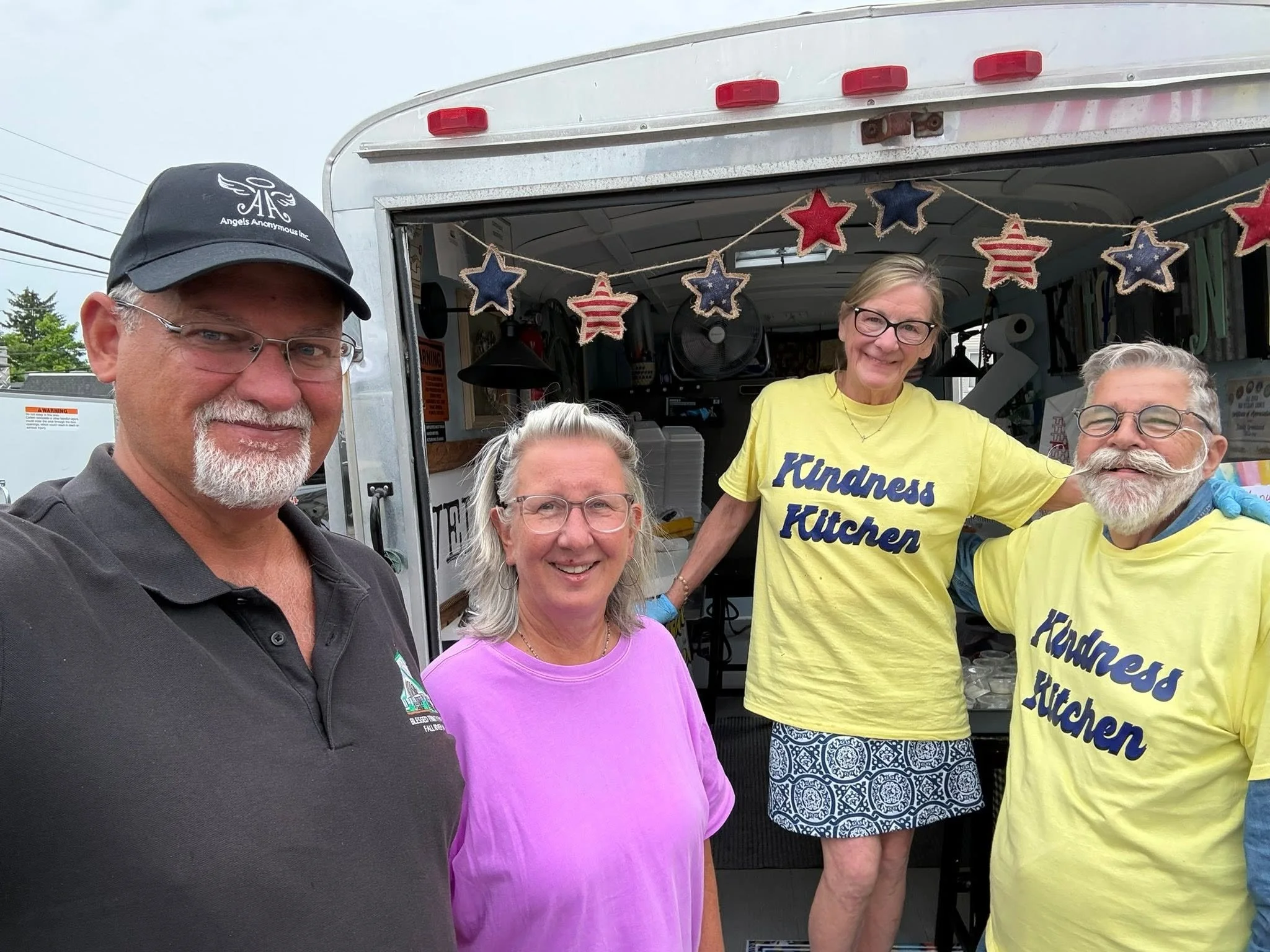 A group of five people standing in front of a food truck decorated with red, white, and blue stars and stripes. The group includes men and women of various ages, smiling at the camera. Two of the women are wearing yellow t-shirts with the words "Kind