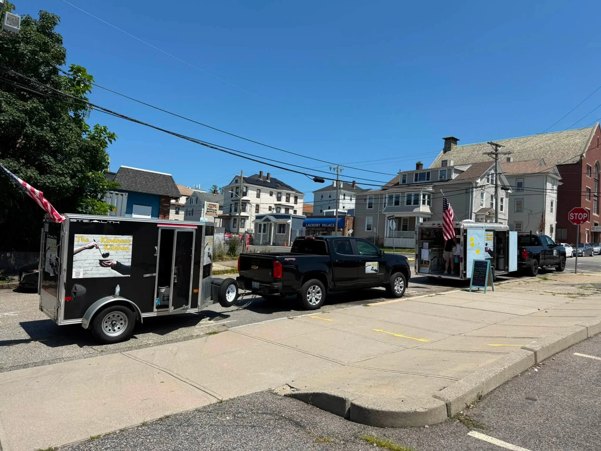 Street view with parked cars, food truck named 'The Kindness Kitchen', and residential buildings with blue sky overhead.