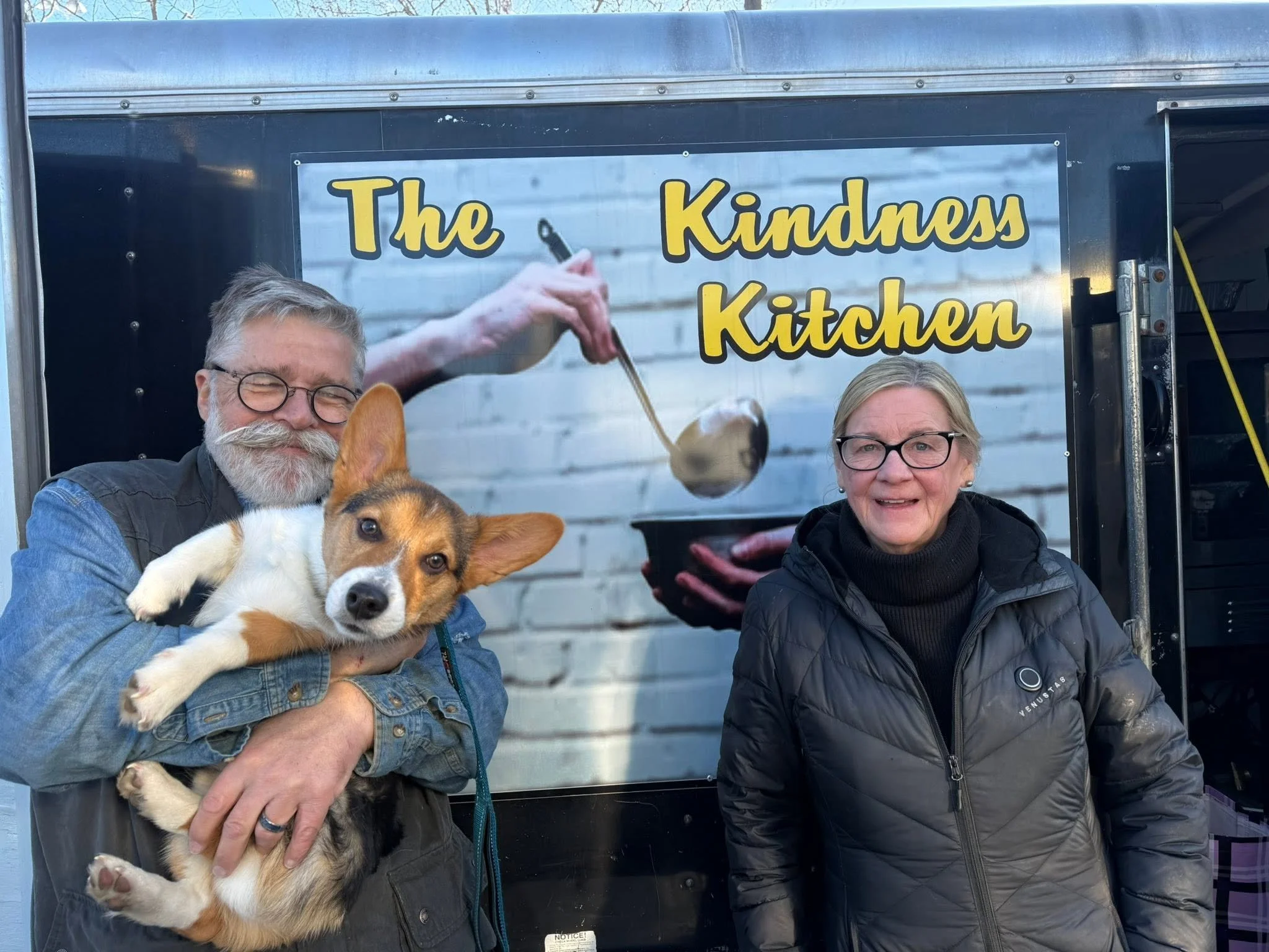 Two people, a man with gray hair, glasses, and a beard, and a woman with short gray hair and glasses, stand outdoors in front of a sign that reads 'The Kindness Kitchen.' The man is holding a dog with a white and brown coat. The dog is lying on its back with its paws relaxed. The woman is smiling and dressed in a black jacket. The background shows a building and the photo appears to be taken on a bright day.