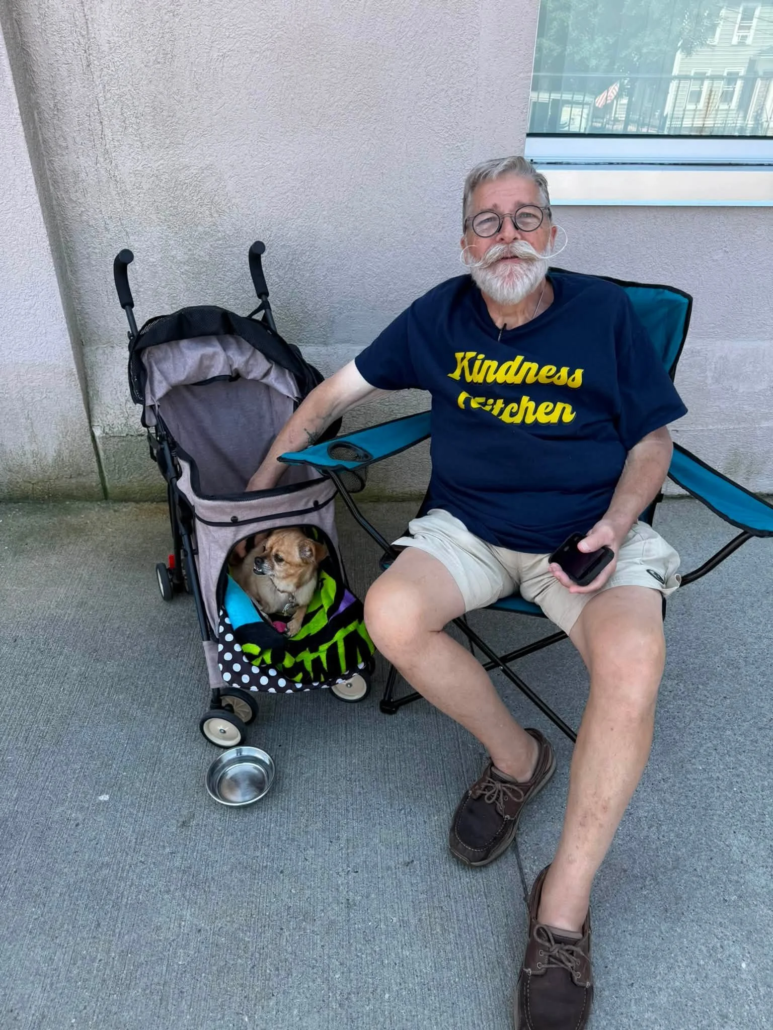 An elderly man with gray hair, beard, and glasses sitting in a blue camping chair on a sidewalk. He is wearing a navy T-shirt with yellow text that reads 'Kindness Kitchen' and beige shorts. His right arm is resting on a stroller with a small dog inside, and he is holding a smartphone in his left hand. There is a metal food or water bowl on the ground nearby.