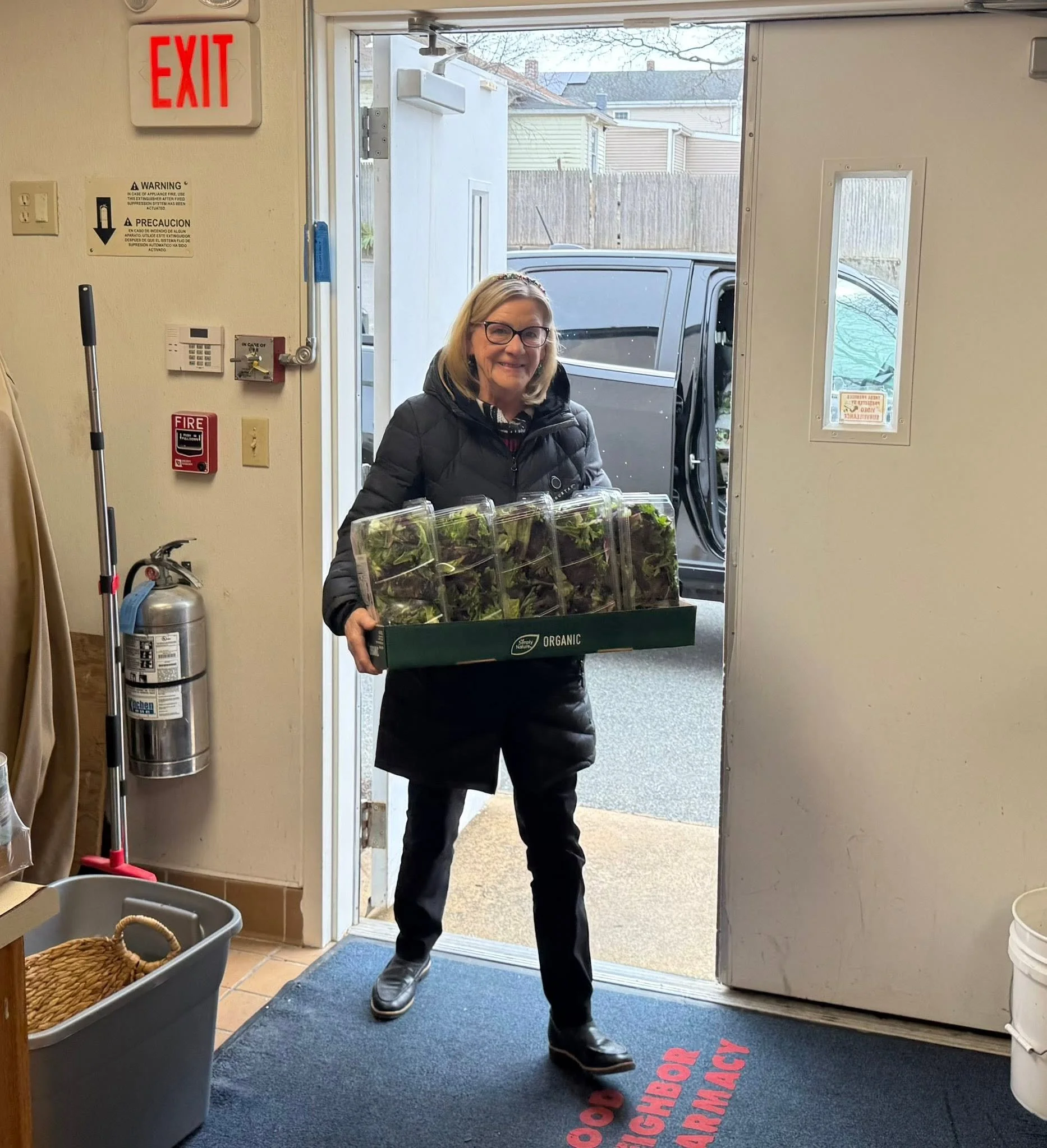 A woman wearing glasses and a black jacket stands inside an entryway, holding a box of organic salad greens. The door behind her is open to an outdoor area with a parked vehicle and wooden houses in the background. Inside, there are signs, a fire alarm, a broom, and a trash can.