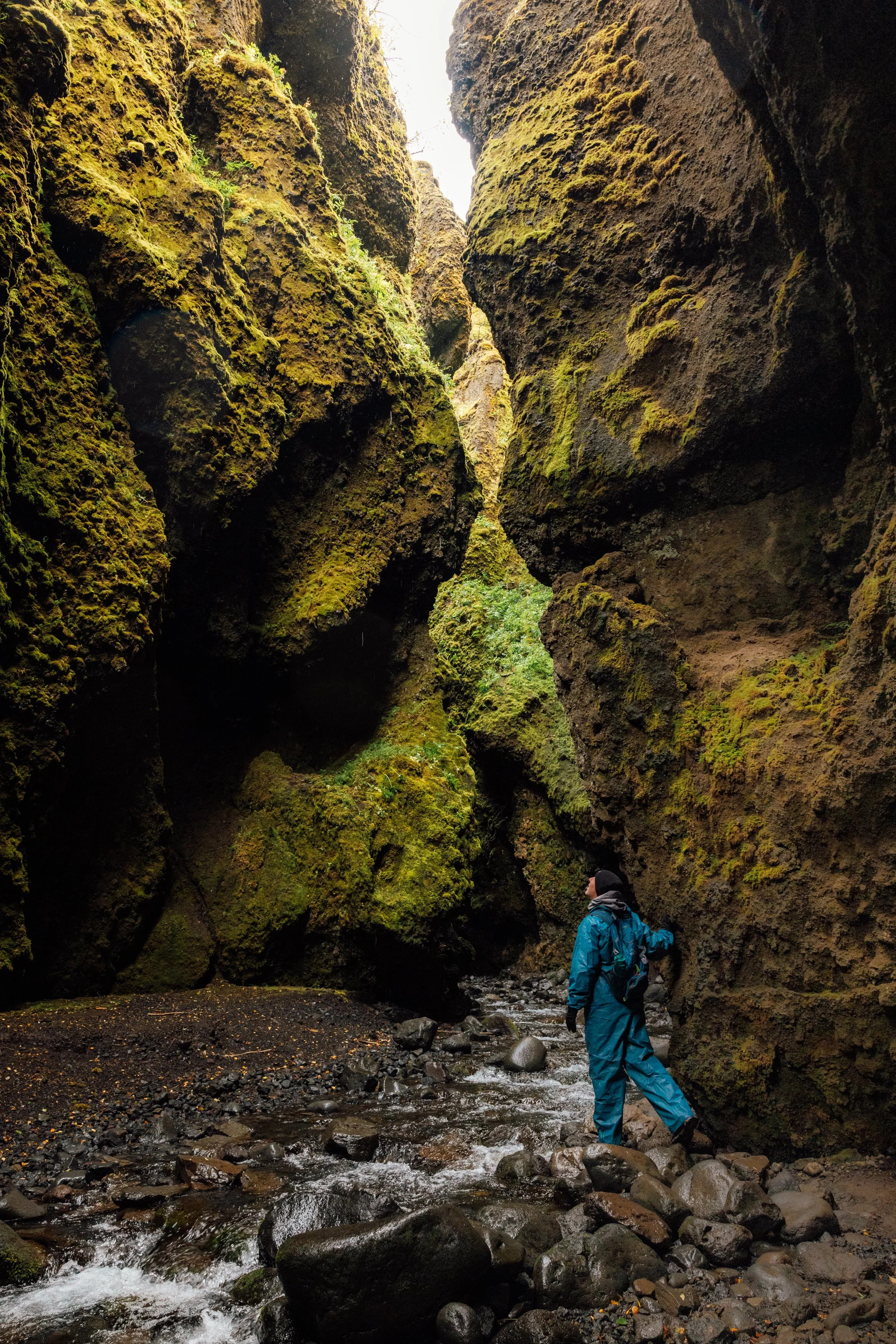 A person dressed in blue waterproof gear walks along a rocky stream in a deep canyon with moss-covered cliffs on either side.