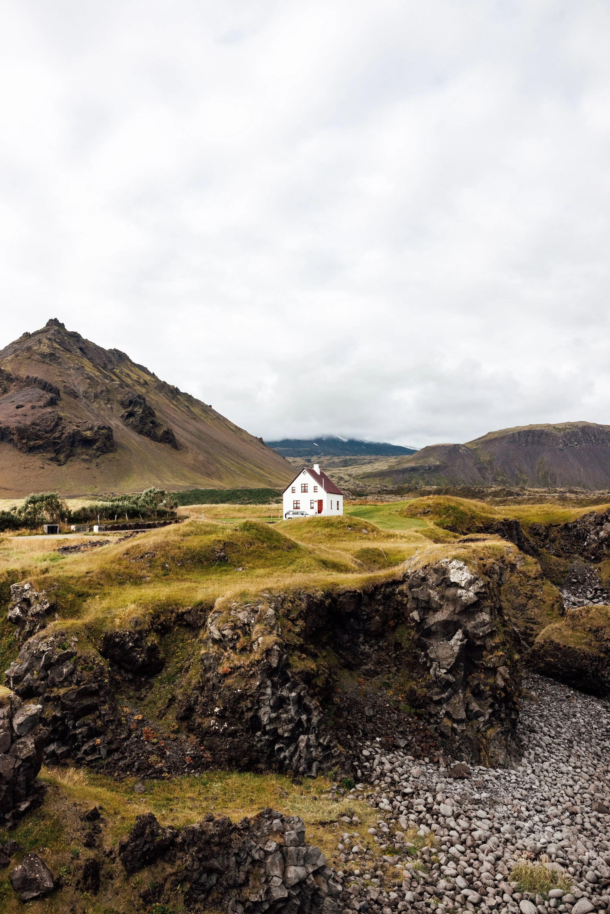 A white house with a red roof in a landscape of green rolling hills, black volcanic mountains, and a cloudy sky.