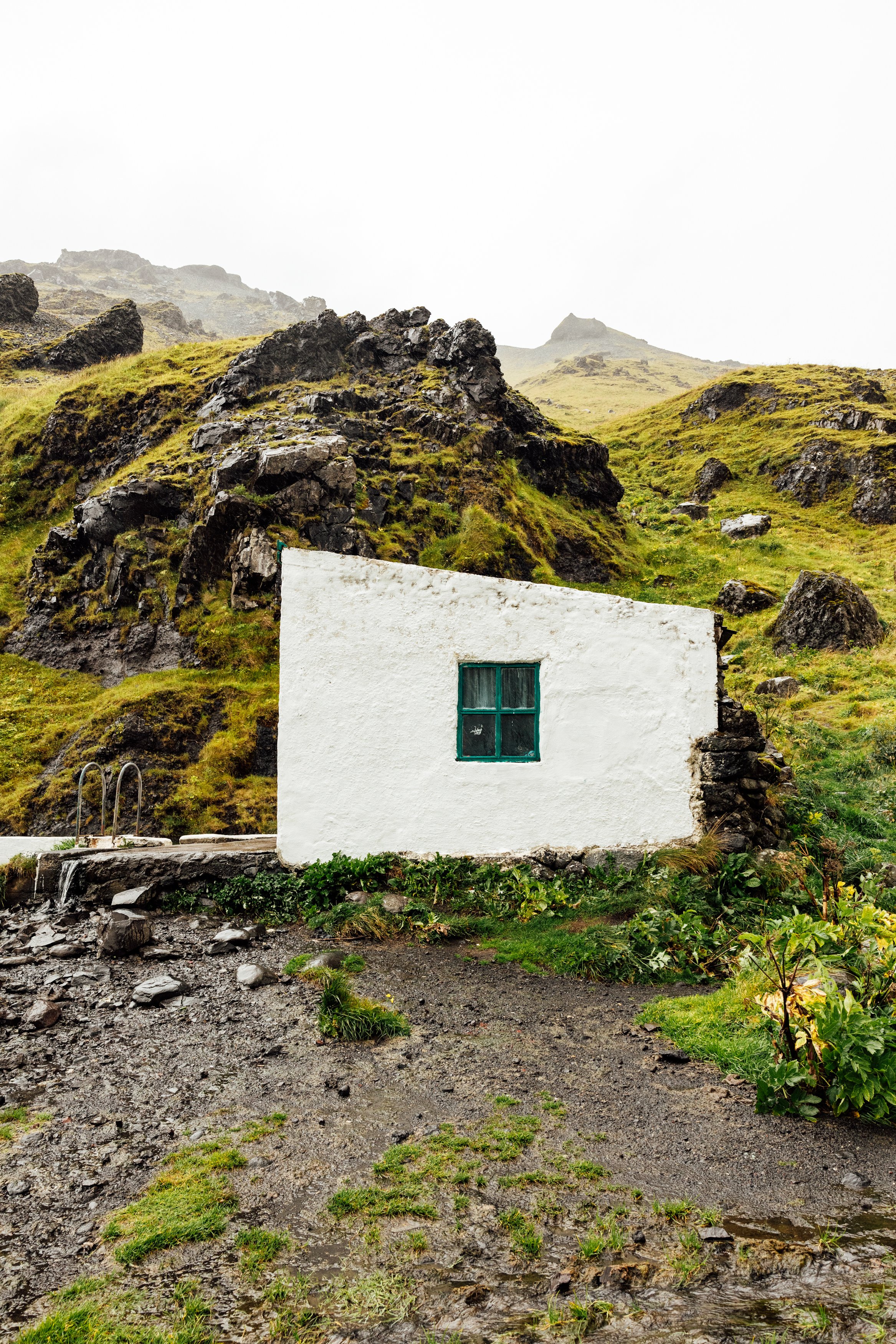 A small white house with a green-framed window set against a grassy hillside with rocks and moss, on a cloudy day.