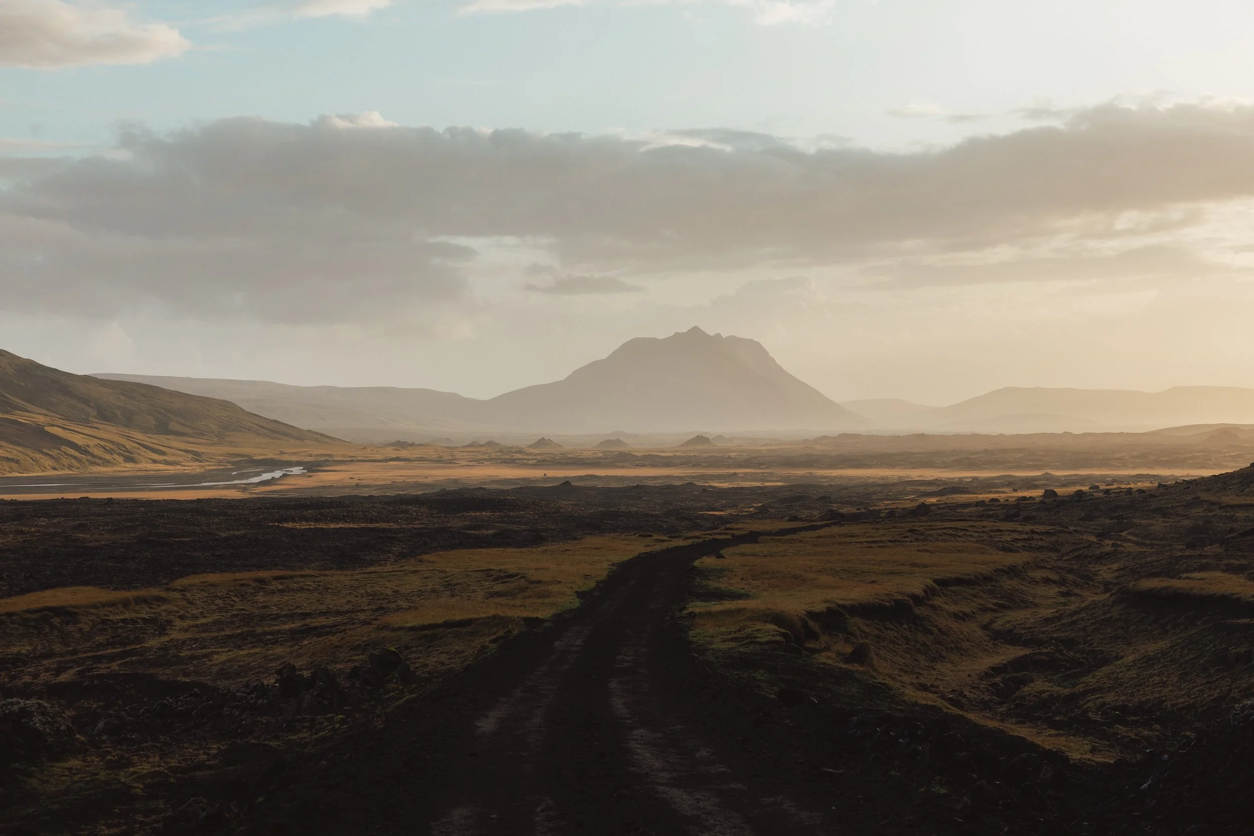 A dirt trail winds through a grassy, volcanic landscape with mountains in the distance under a cloudy sky.