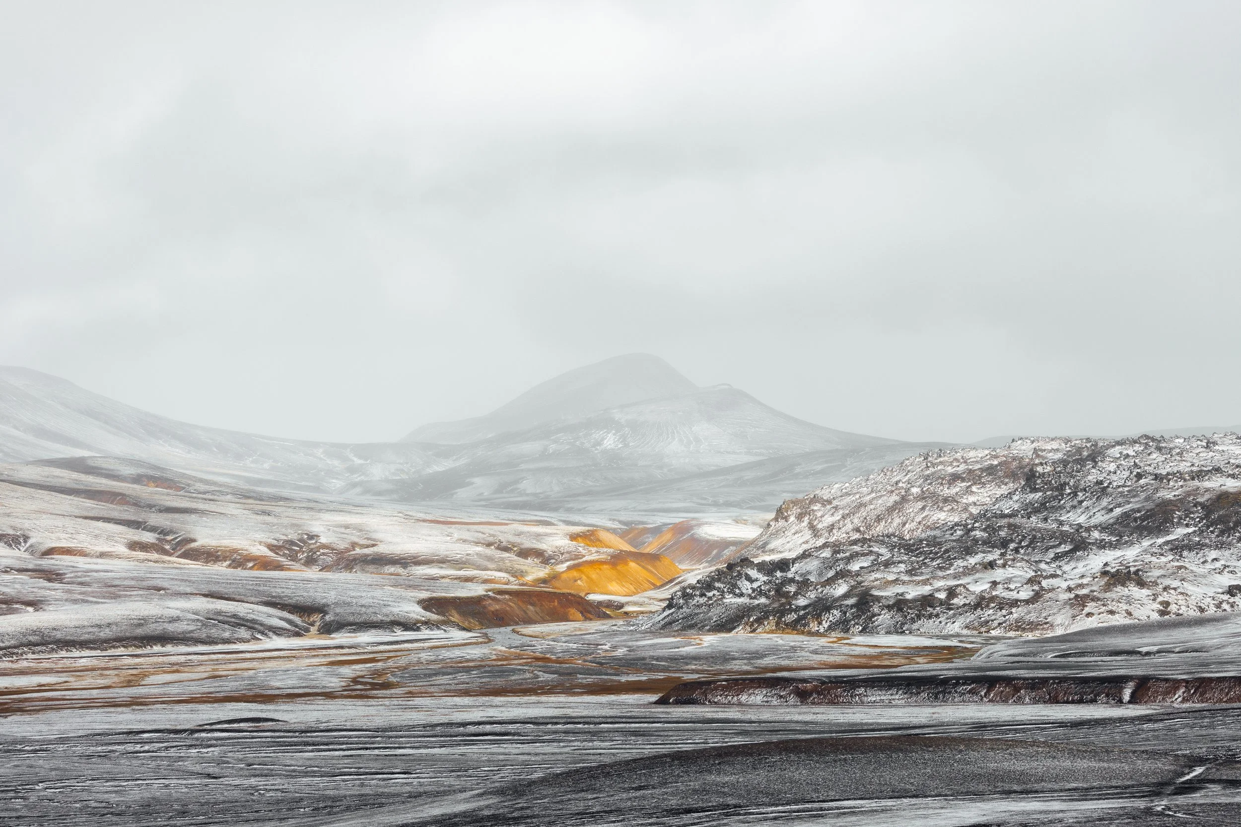 A vast volcanic landscape with snow-covered terrain, colorful geothermal features, and distant mountain peaks under a cloudy sky.
