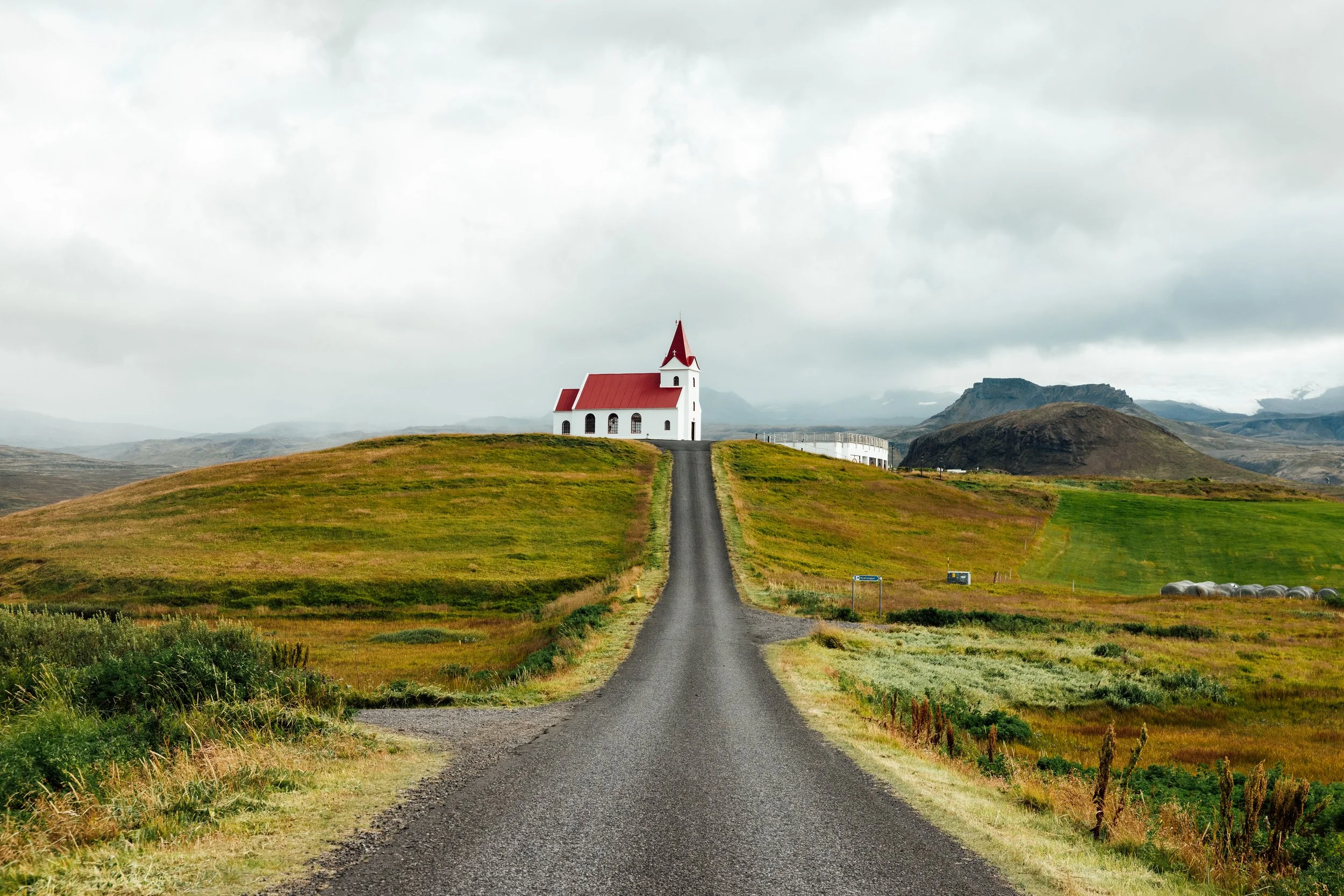 A church with a red roof and steeple located on a hilltop, reached via a long, straight gravel road, surrounded by green fields and rolling hills under a cloudy sky.