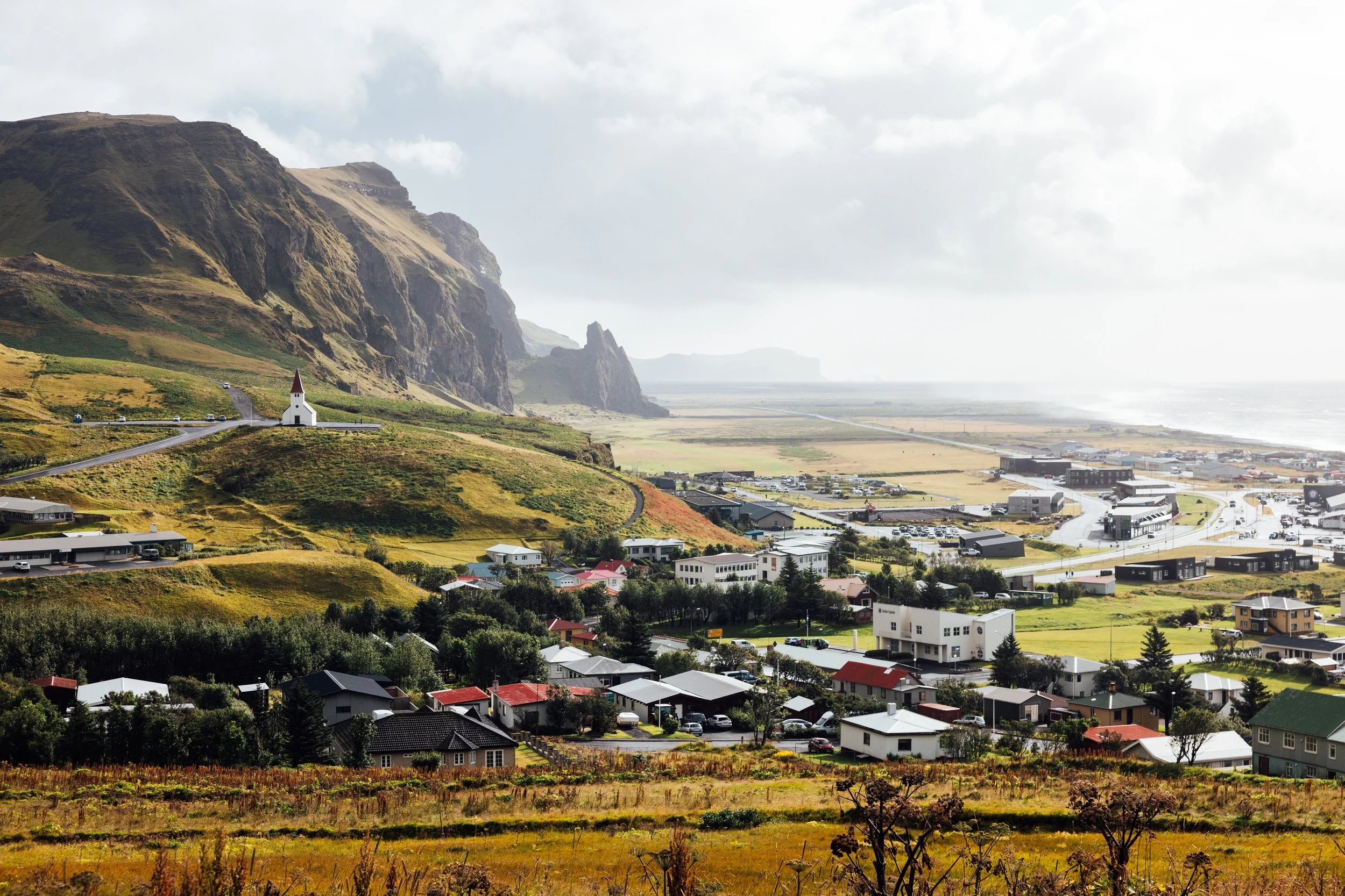 A coastal town with a mix of houses, a small church with a red roof on a hill, green fields, and a mountain range in the background, near the ocean.