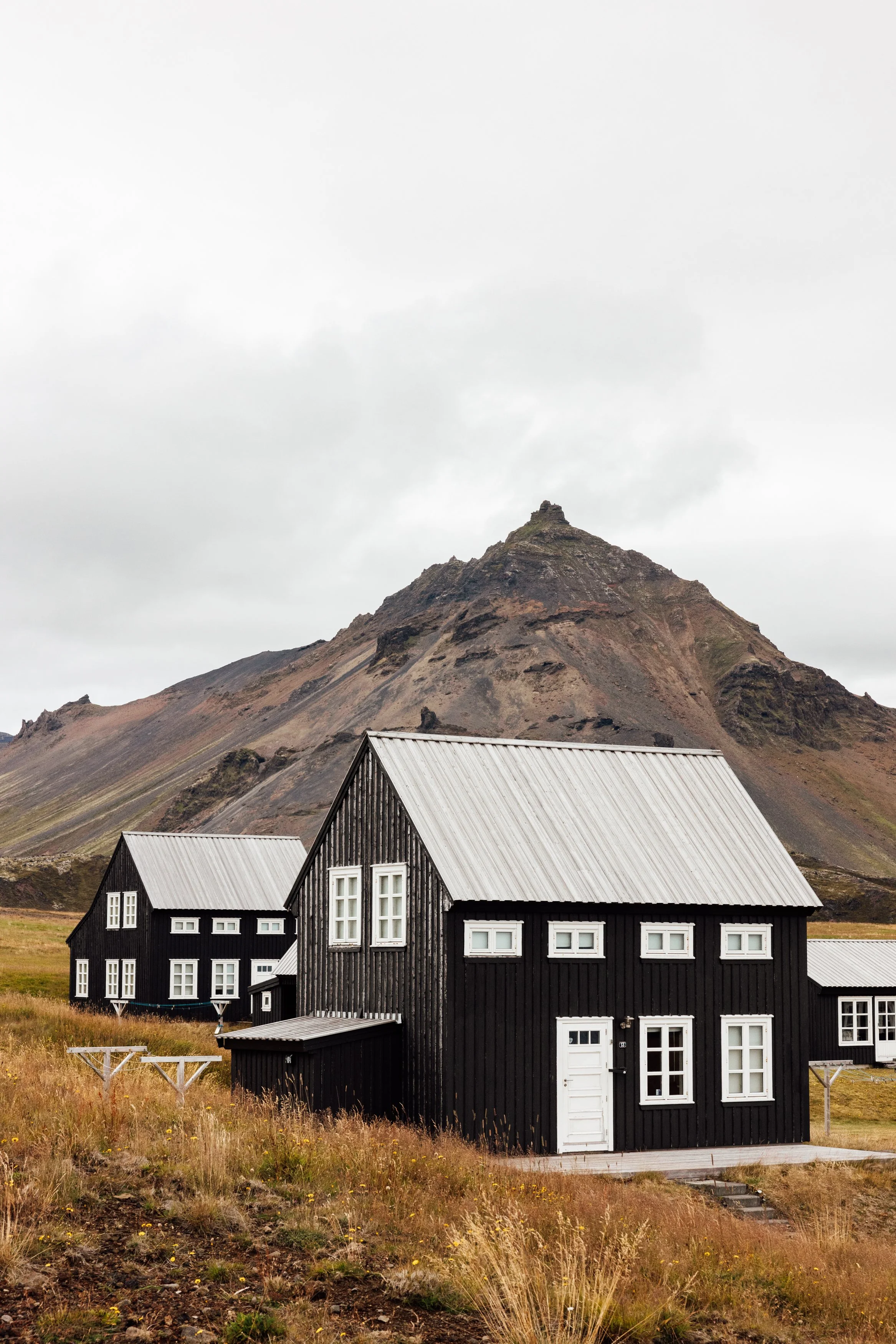 Black wooden houses with white window frames and metal roofs set against a rugged mountain with overcast sky.