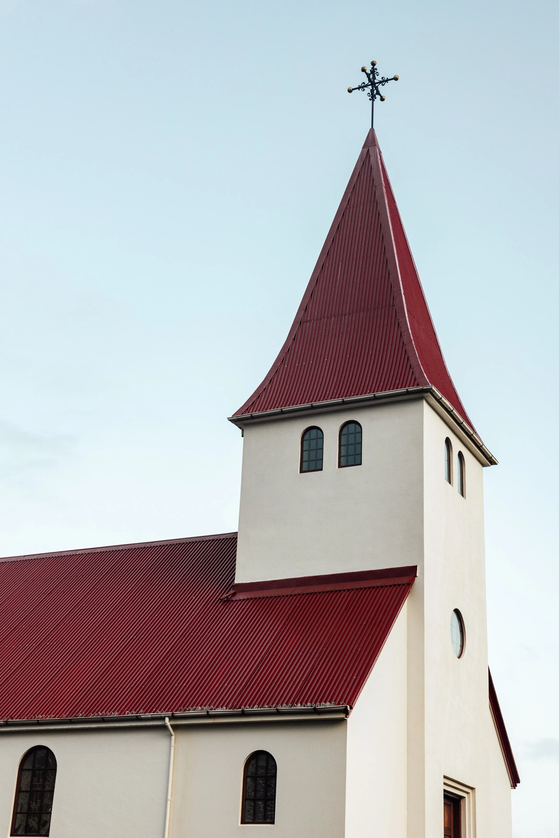 A church with a red, steeply pointed roof and a white exterior, featuring small arched windows and a cross on top of the steeple.