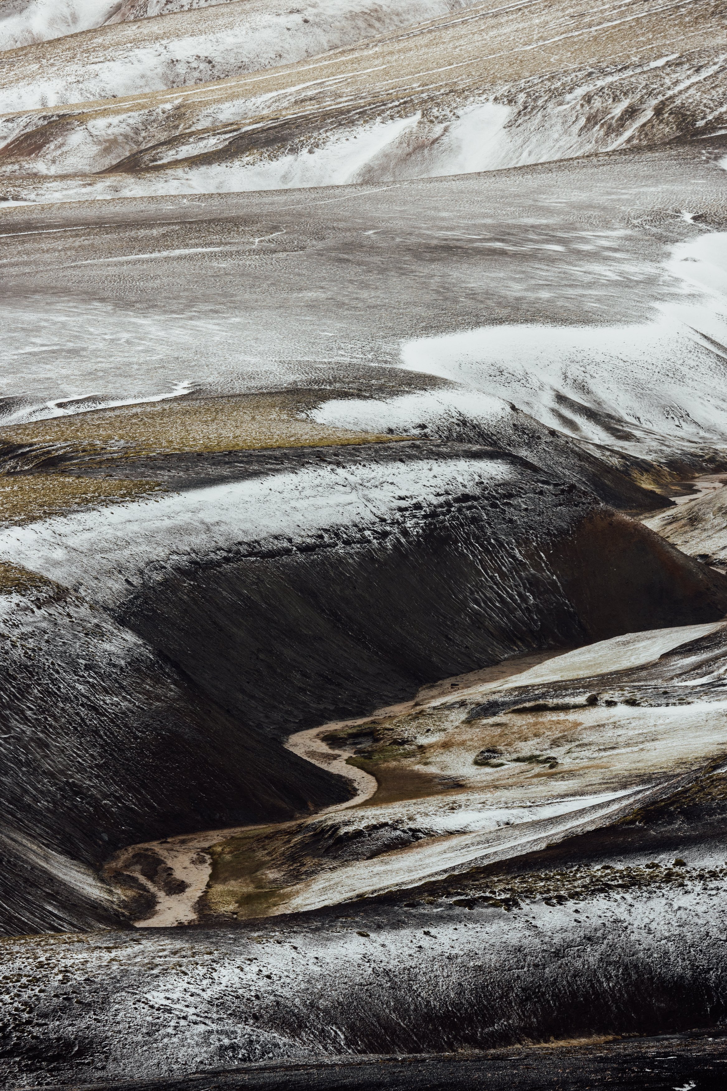 A rugged, snow-dusted landscape of mountains with deep valleys and winding streams, featuring dark exposed rock and patches of snow and snow-covered slopes.