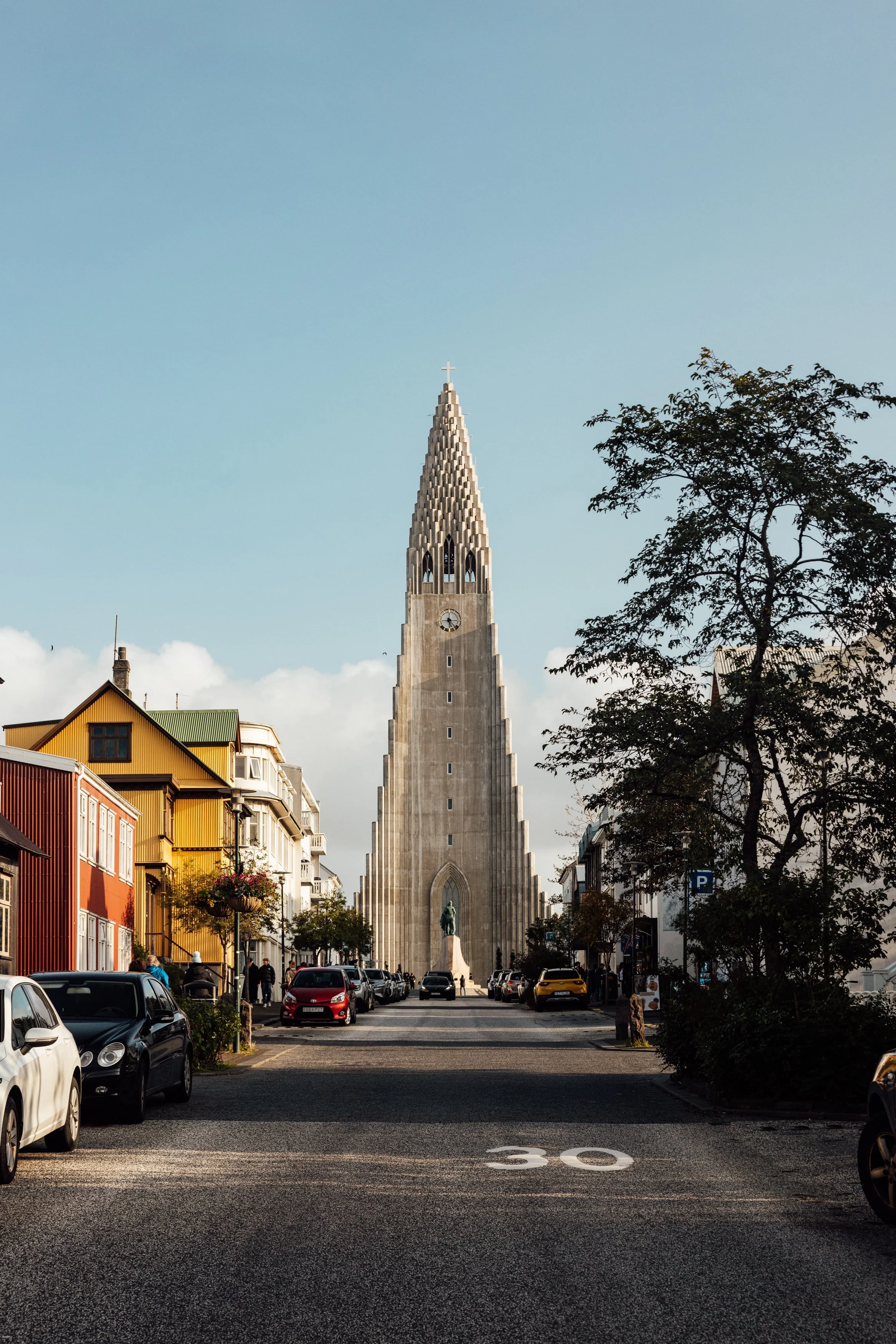 Street view of Hallgrímskirkja church in Reykjavik, Iceland, with cars parked along the street, colorful buildings on the left, trees on the right, and a blue sky with some clouds.