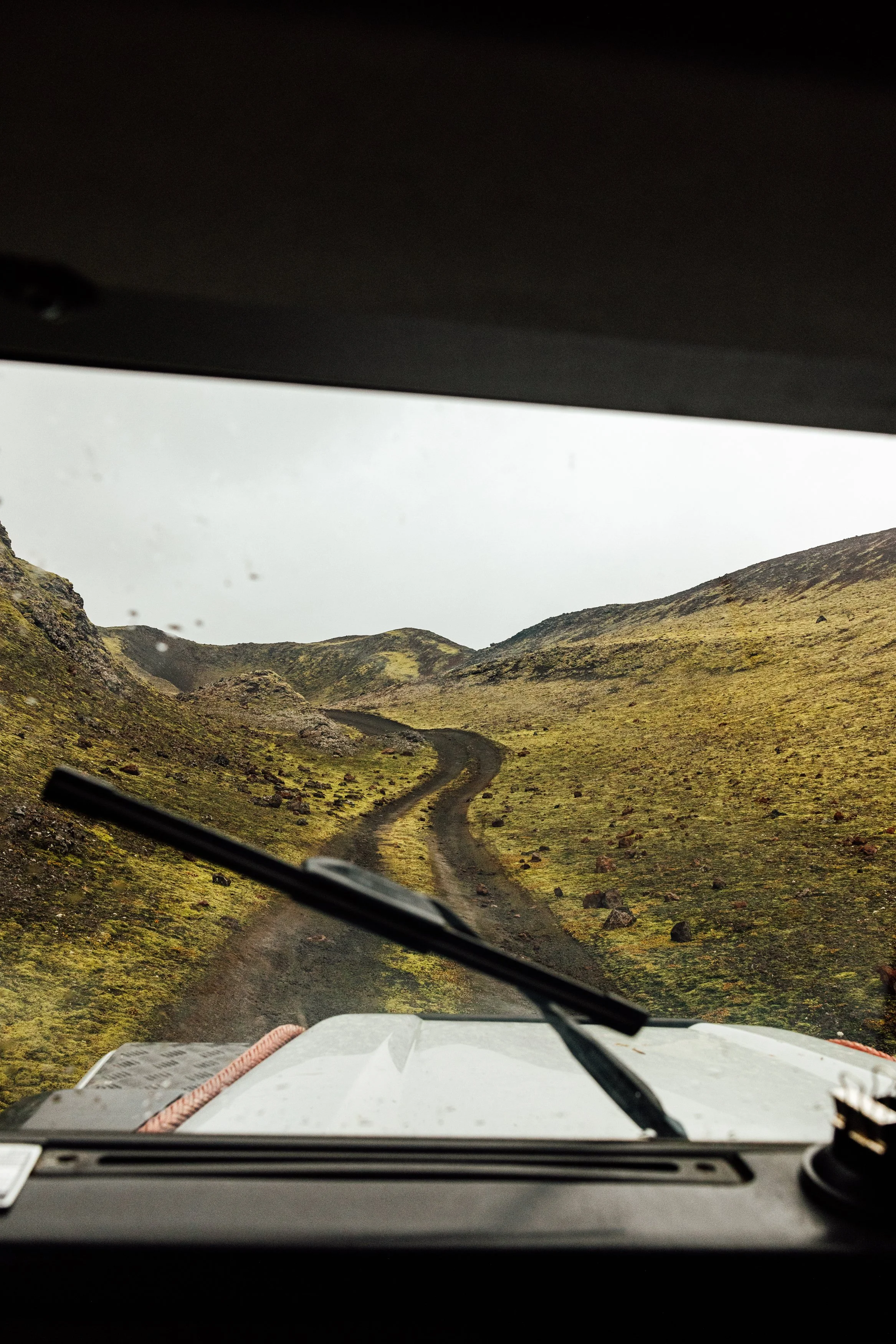 Dirt road winding through moss-covered, rocky hills under an overcast sky, viewed from inside a vehicle.