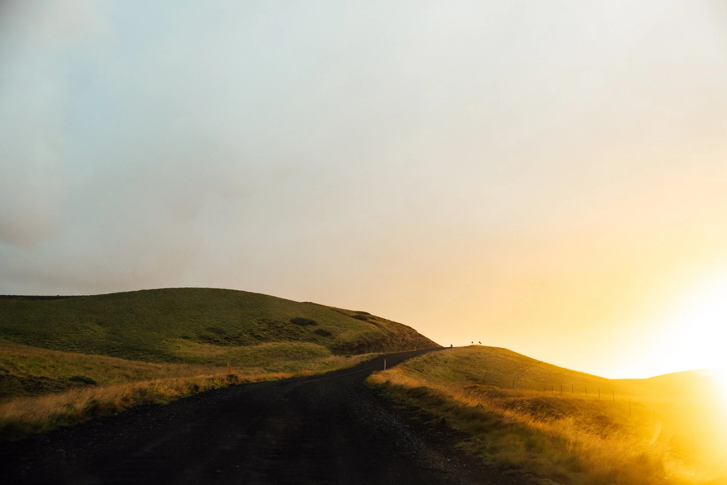 A winding dirt road through green rolling hills at sunset with a bright sun on the horizon.