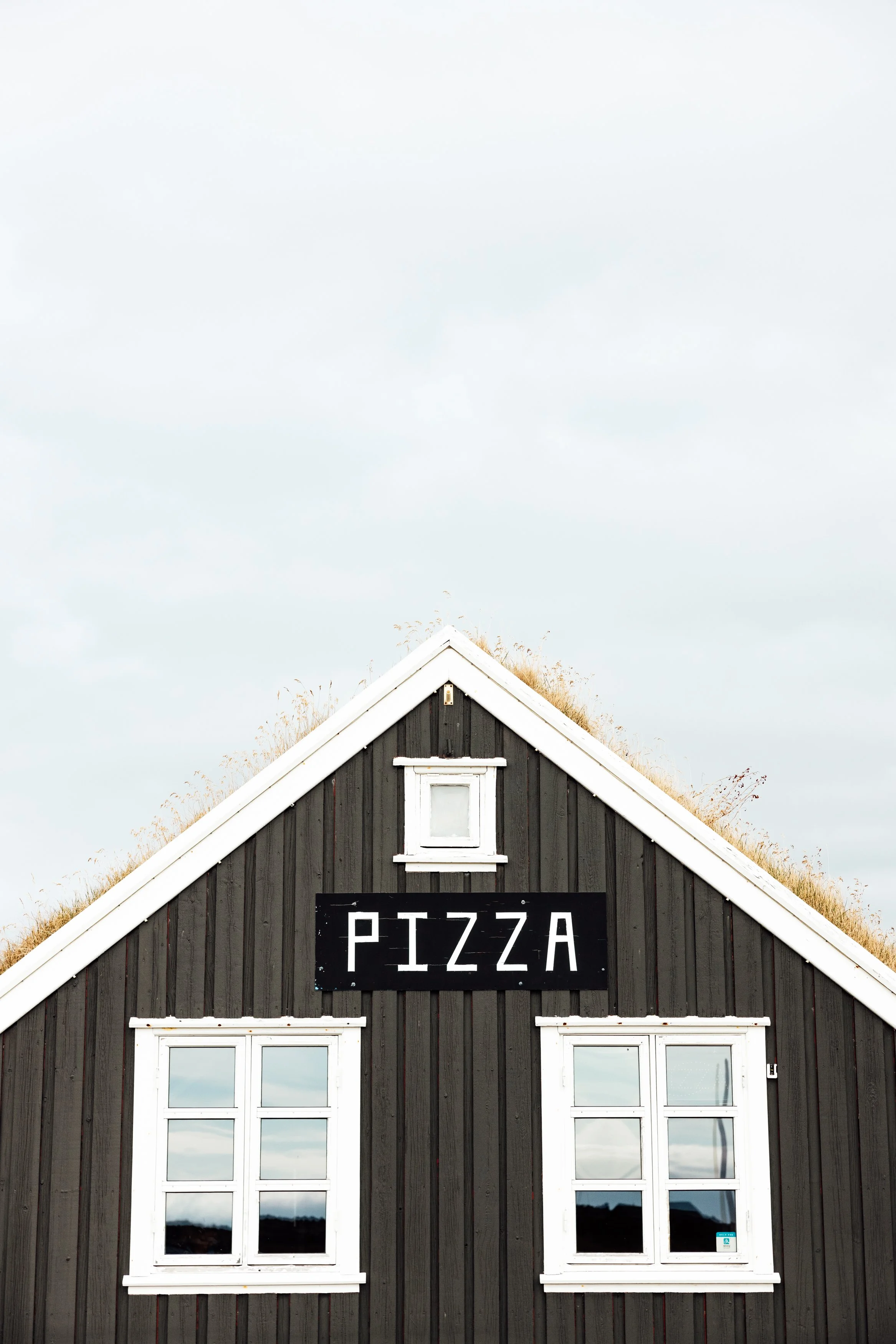 A black wooden building with white trim and a pitched roof, featuring a black sign that reads "PIZZA" in white letters.