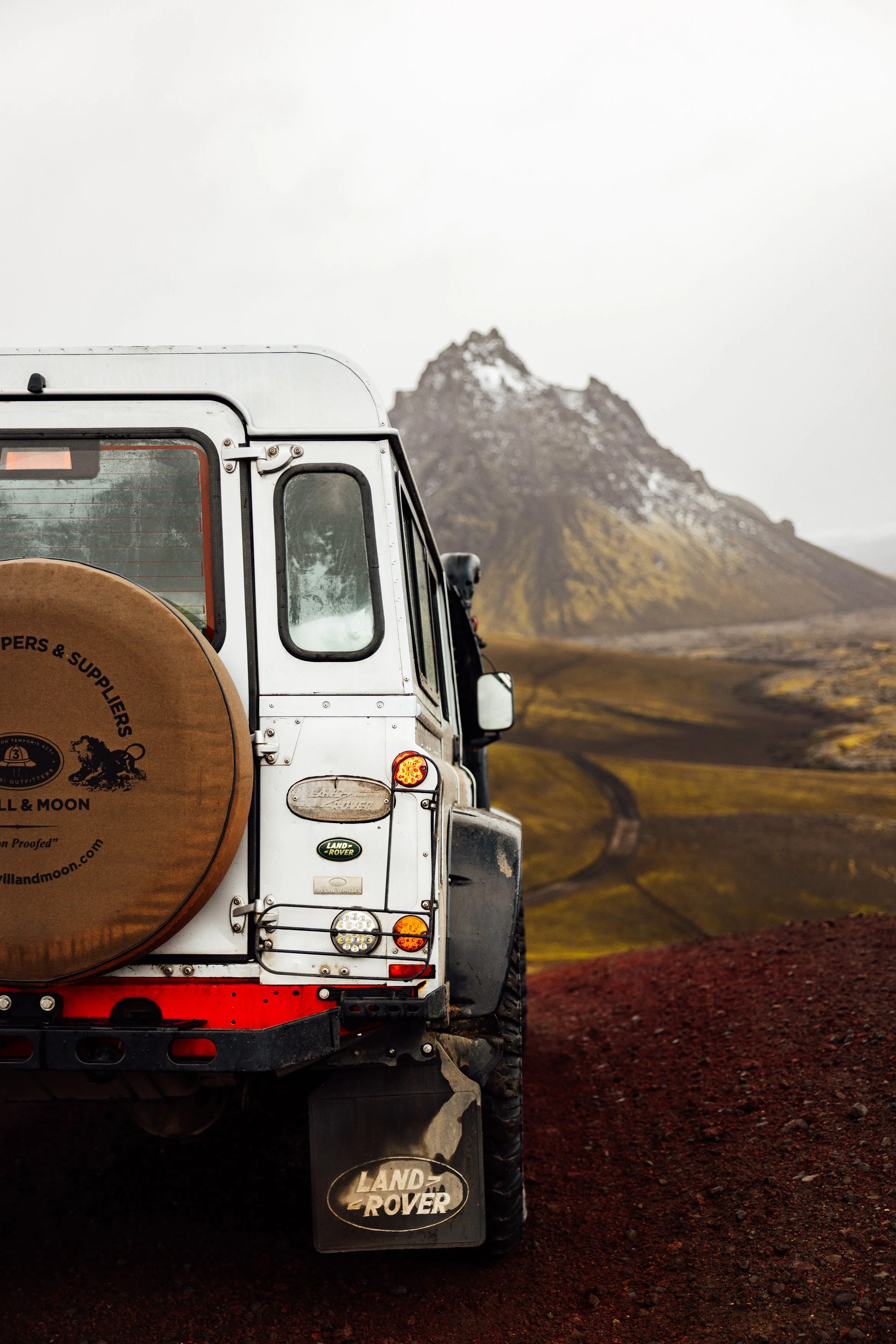 Back of a white Land Rover vehicle on a dirt road with a mountain in the background.